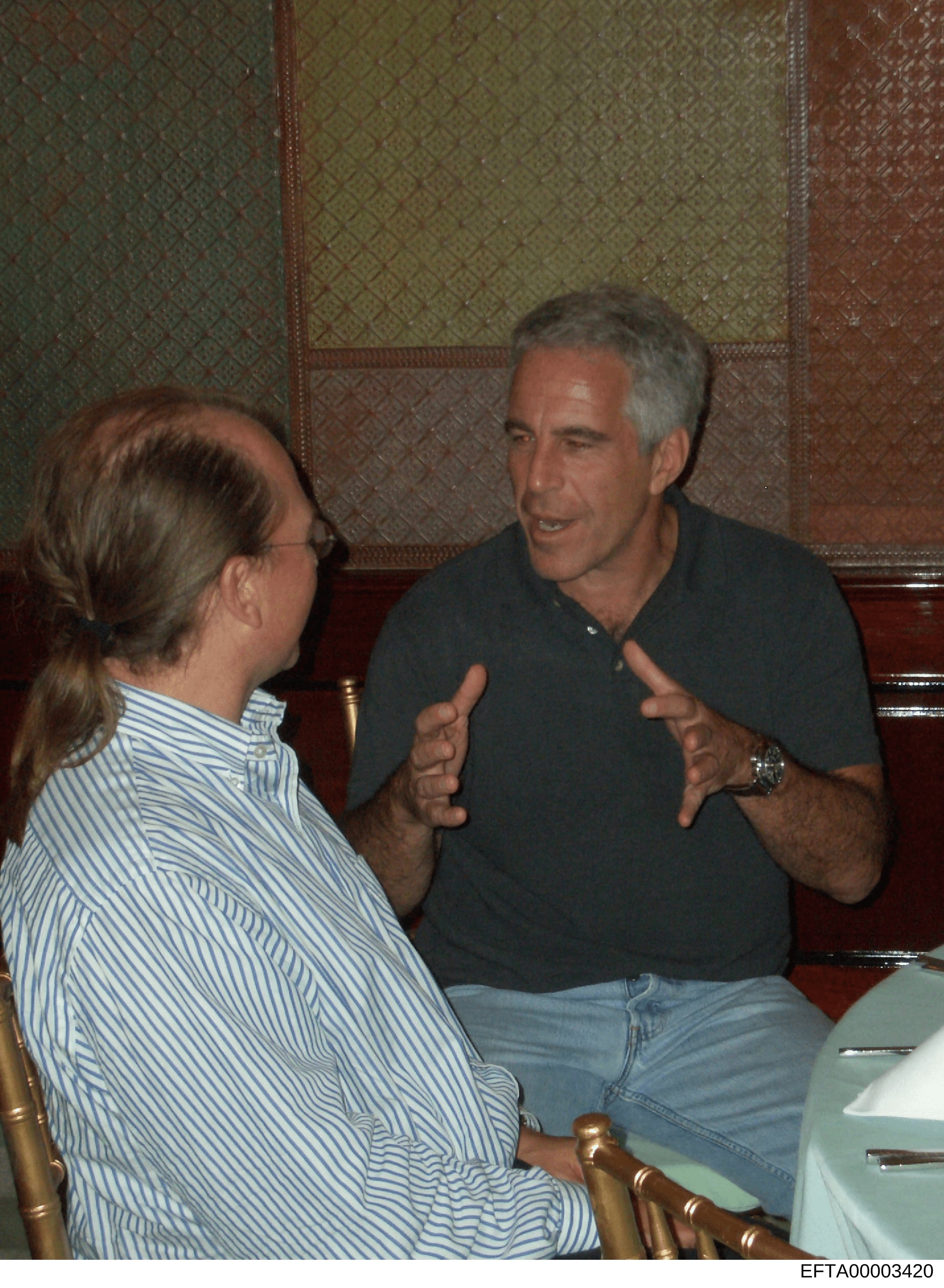 A color photograph showing two people in conversation in an interior room with decorative wallpapered walls. The image appears to be from the 1990s based on the photo quality and style. One individual is seated wearing a blue and white striped shirt,