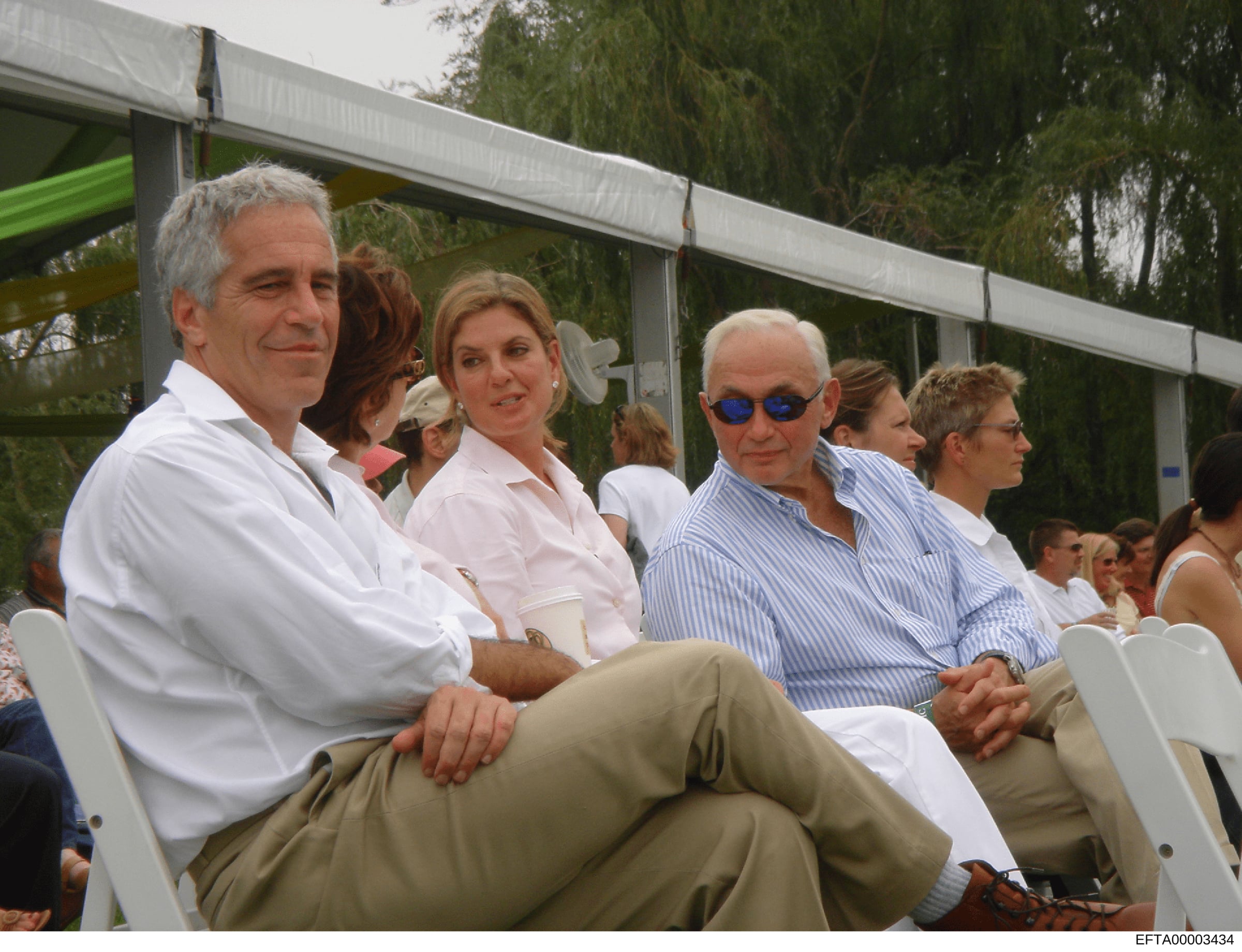 This photograph shows a group of people seated at an outdoor event under a covered pavilion structure. The image captures multiple attendees in casual-formal attire, with trees visible in the background. The photo is dated and catalogued as evidence 