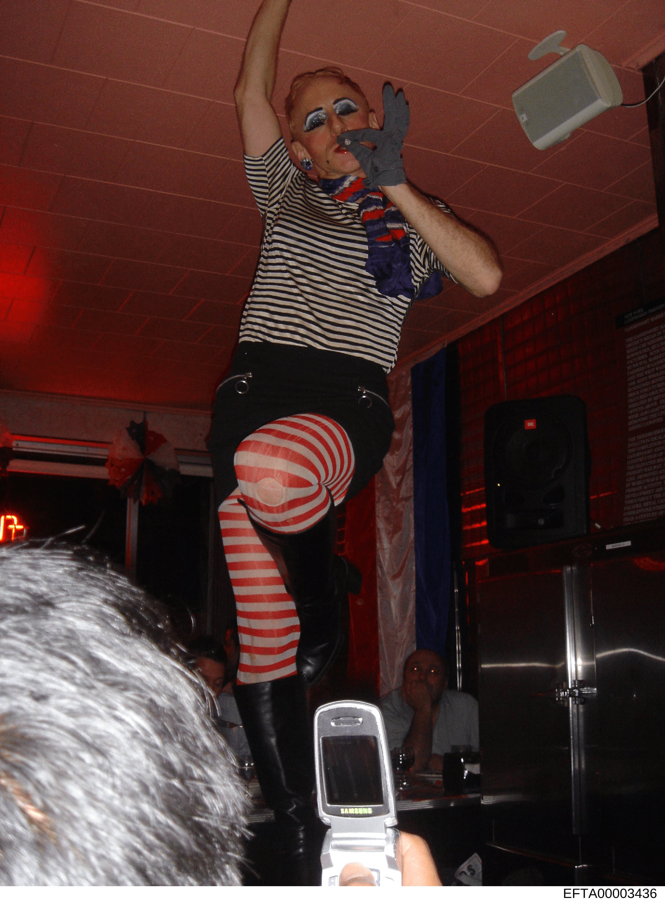 This is a photograph taken at what appears to be an indoor venue or nightclub with red lighting. The image shows a person wearing striped clothing with theatrical makeup and styling. The photo was taken with a flip phone camera (visible Samsung devic