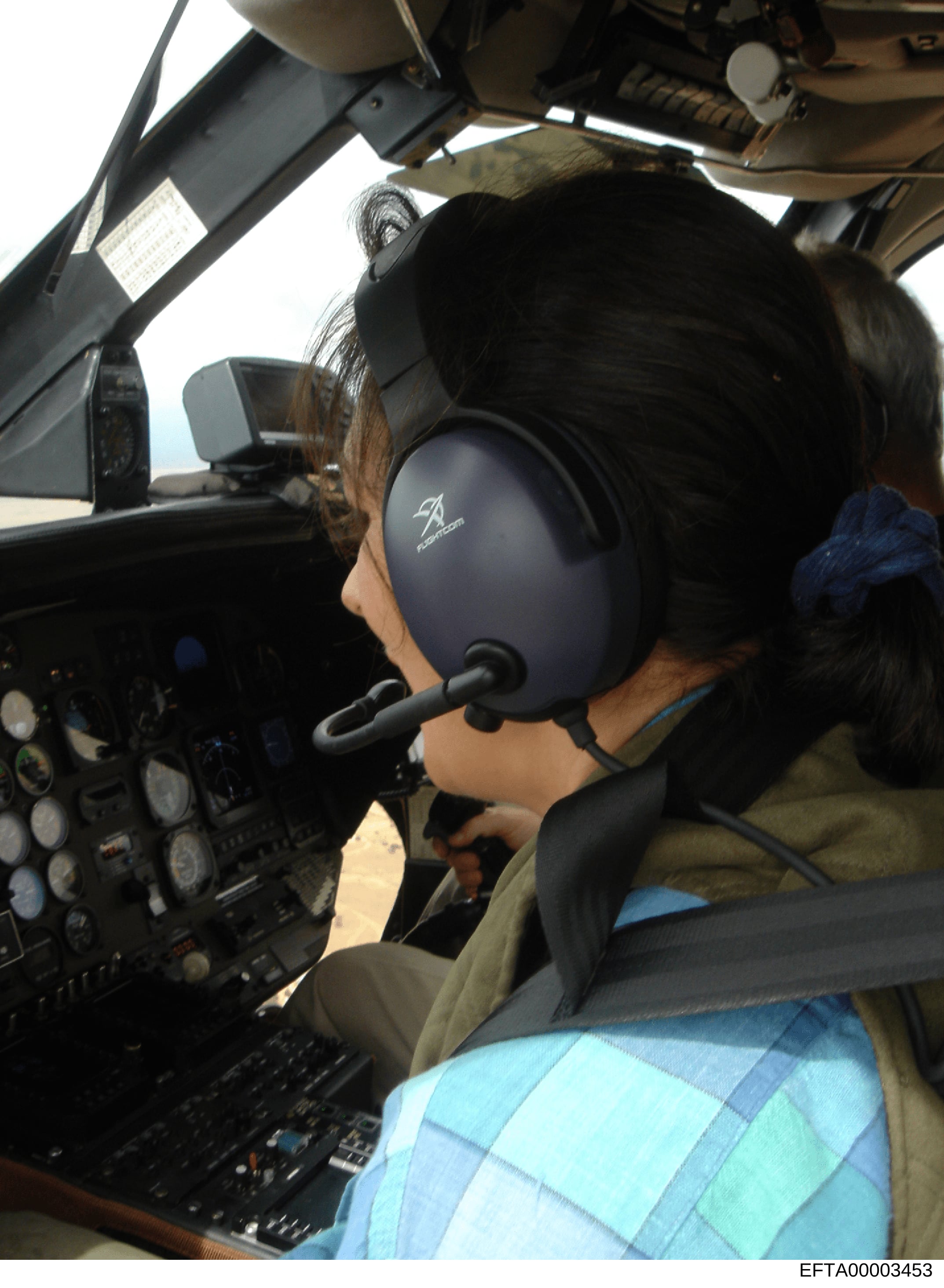 This photograph shows the interior of a helicopter cockpit with a pilot wearing a black headset and olive/tan flight suit seated at the controls. The image captures the instrument panel, overhead systems, and operational environment of what appears t