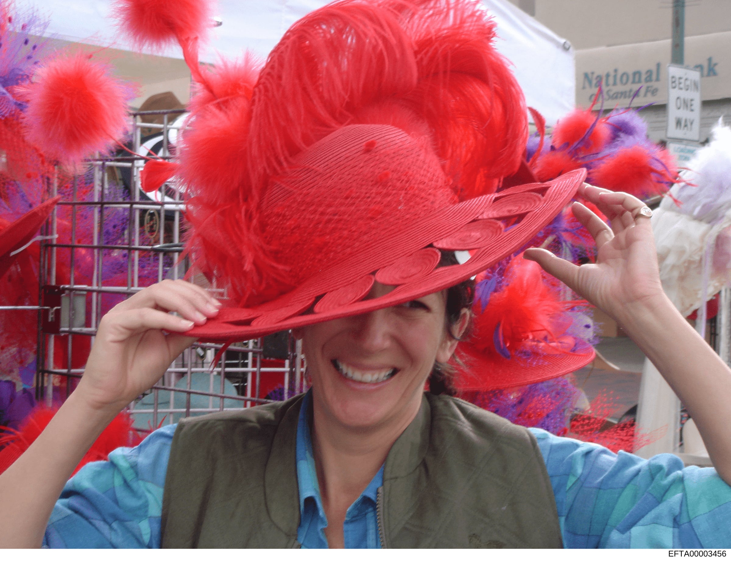 This is a candid photograph of a woman trying on an elaborate red feathered hat with purple accents at what appears to be a public event or street fair. The image shows decorative hats displayed in the background and includes a 'National Bank' sign v