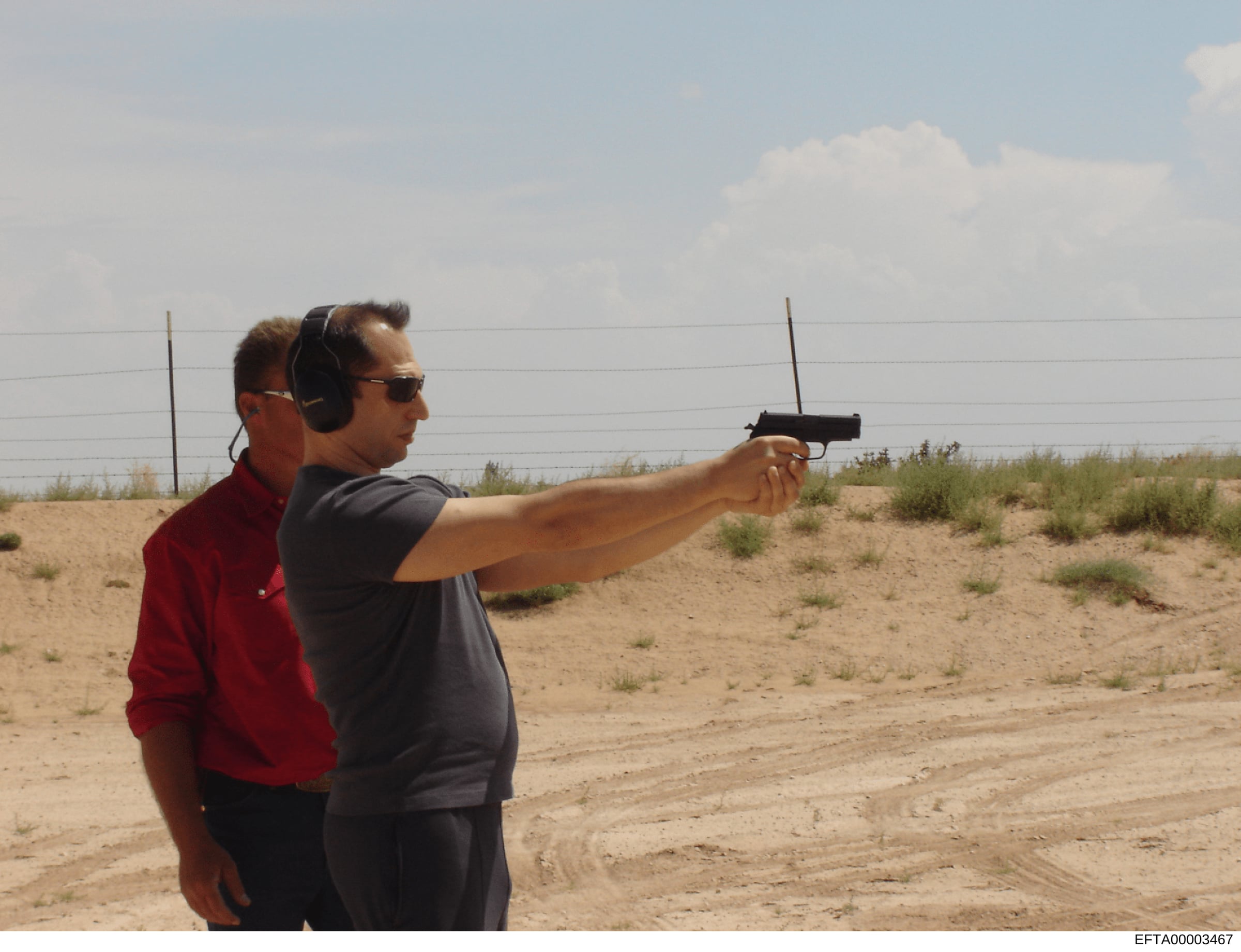 This photograph shows an individual at an outdoor shooting range wearing protective headphones and sunglasses, aiming a handgun downrange. The image appears to be documentary or evidentiary in nature, capturing shooting range activity. The context of