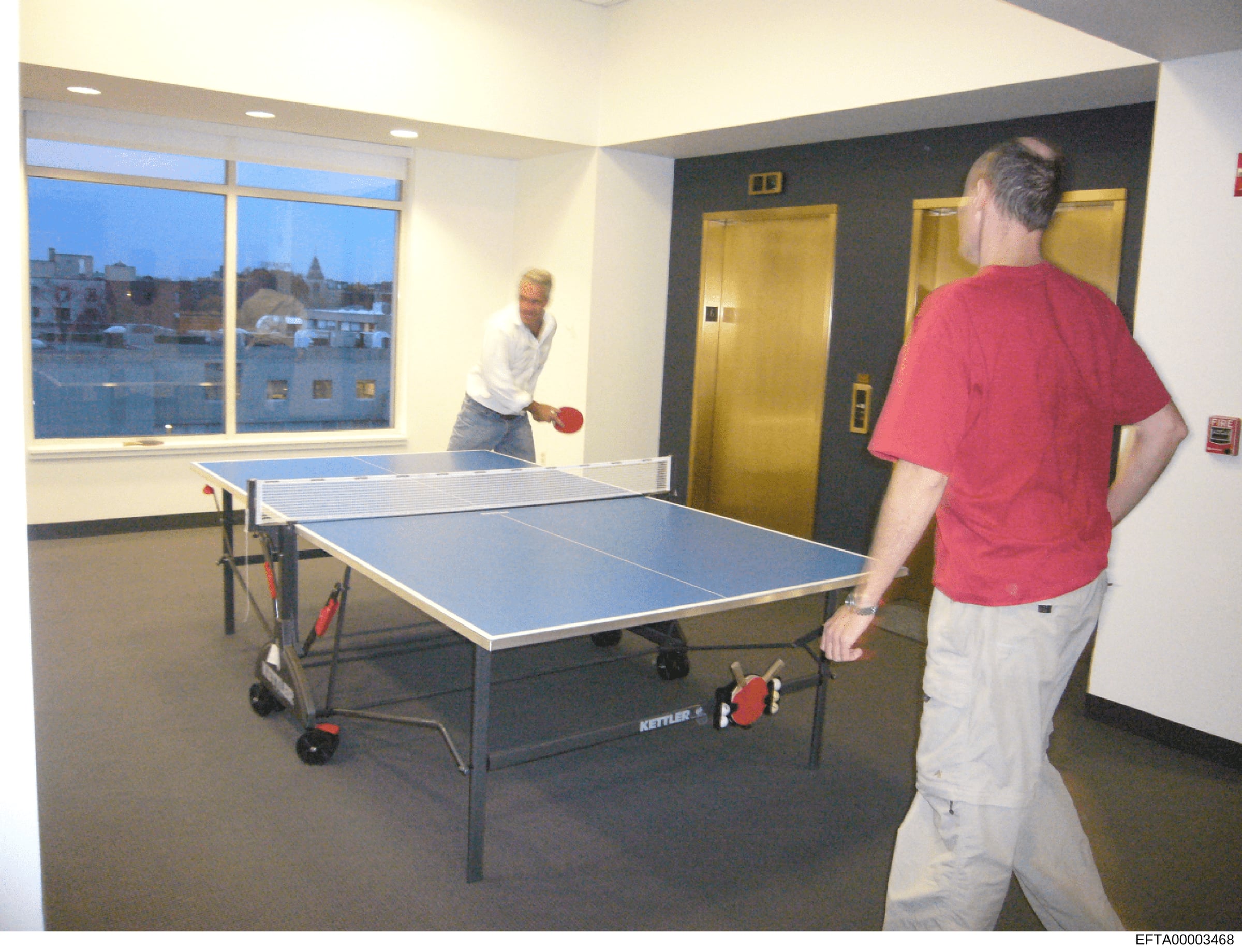 This photograph shows two men playing table tennis in what appears to be a high-rise building's recreation or game room. The room features floor-to-ceiling windows overlooking an urban cityscape at dusk, gold-colored elevators, and modern amenities.