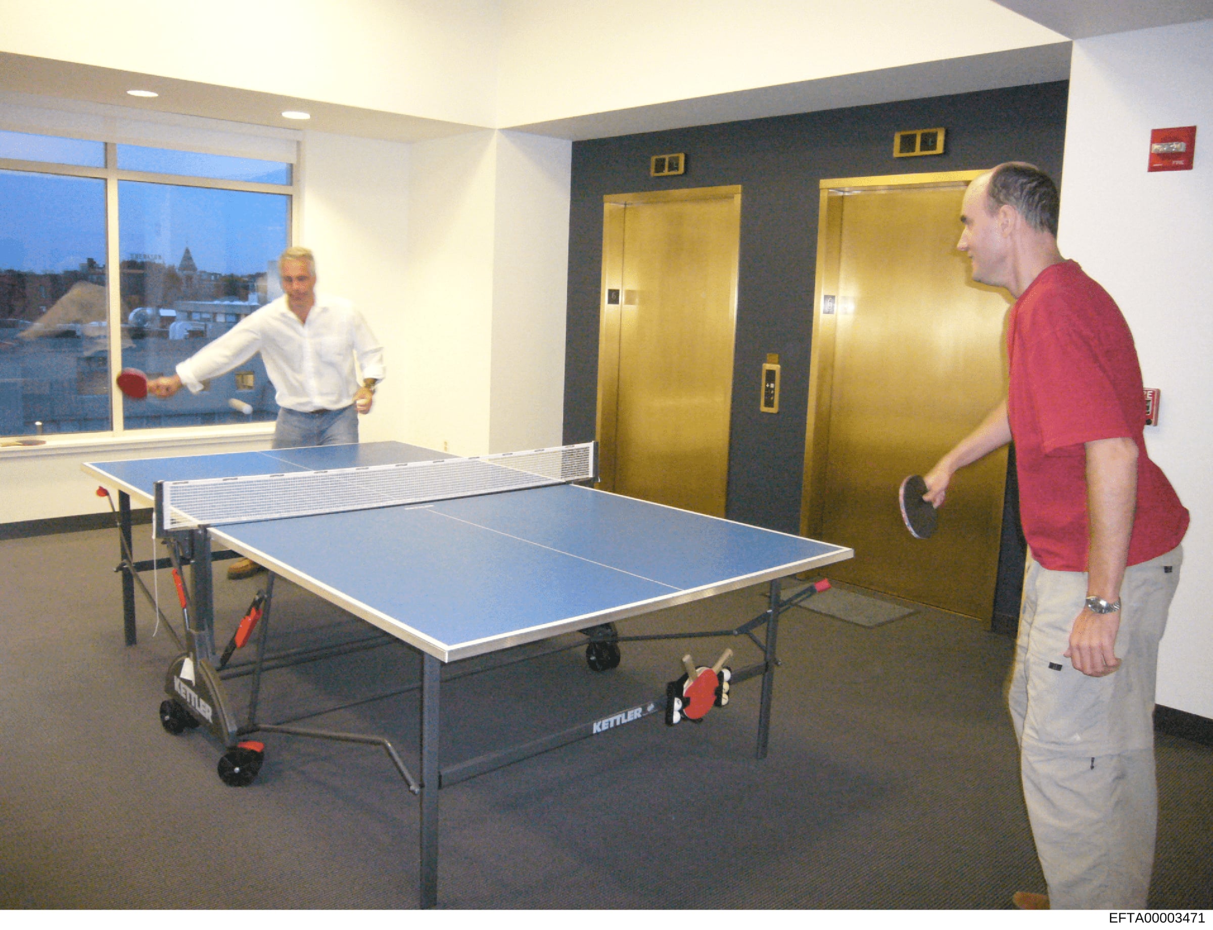 This photograph shows an interior recreational space with a ping pong table and two men engaged in or preparing for a game. The room features large windows overlooking a cityscape, gold-finished elevators, and modern amenities. The photograph appears
