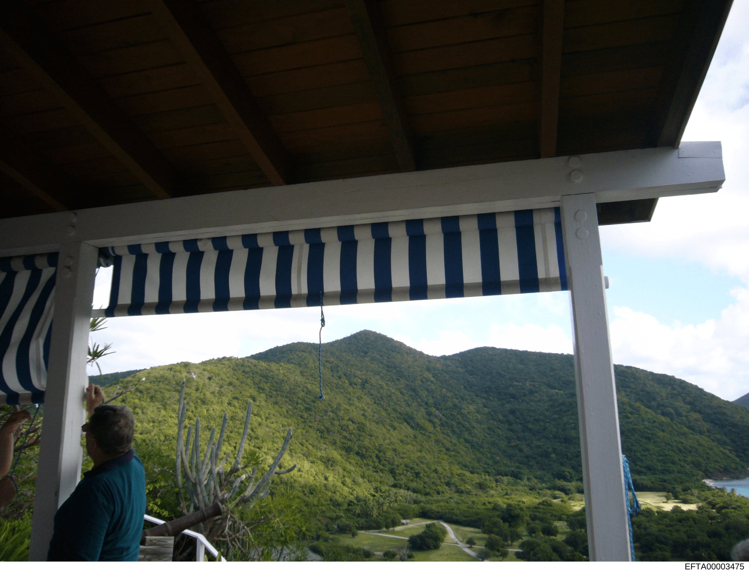 This photograph shows a covered terrace or pergola structure with blue and white striped awning, overlooking a lush mountainous landscape. The image appears to be documentation of a property, possibly one of Epstein's residences, showing the exterior