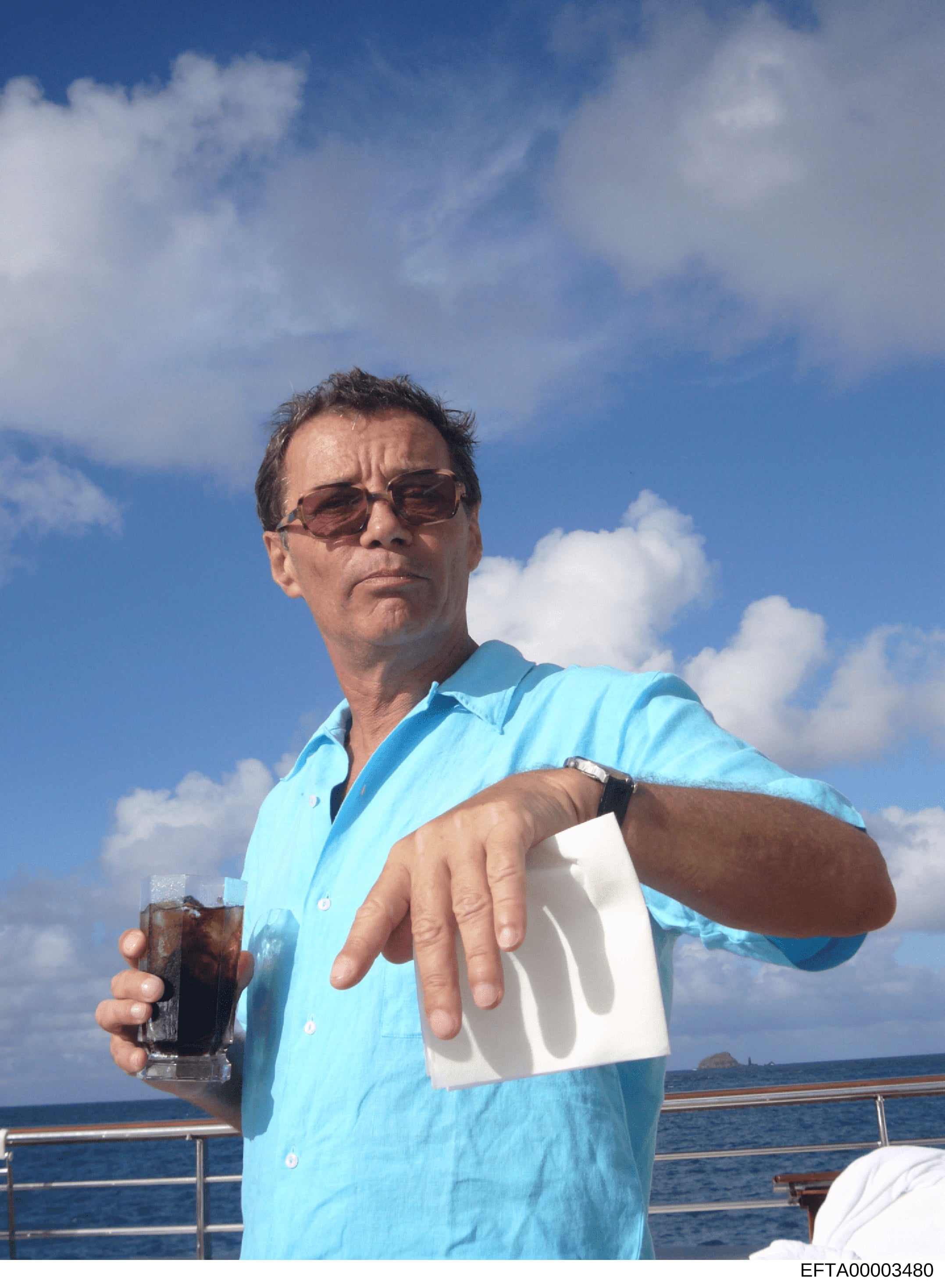 This is a color photograph showing a middle-aged man on what appears to be a yacht or large vessel, holding a dark beverage in one hand and a white document or paper in the other. The photo was taken during daytime with clear blue sky and ocean visib