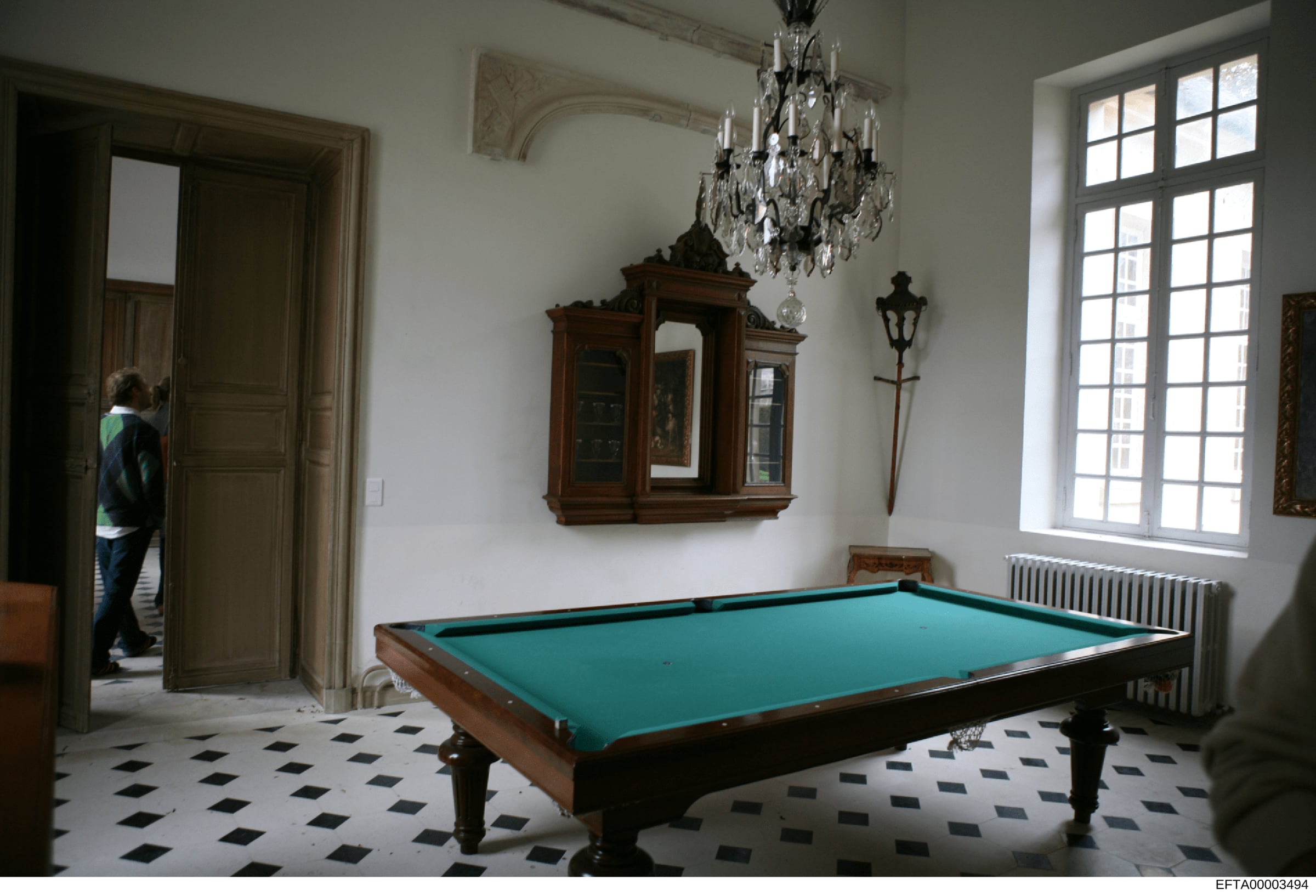 This is a photograph of an elegant interior space featuring a billiards table with teal felt, ornate European-style furnishings, a crystal chandelier, and period architectural details. The room displays high-end residential décor consistent with prop
