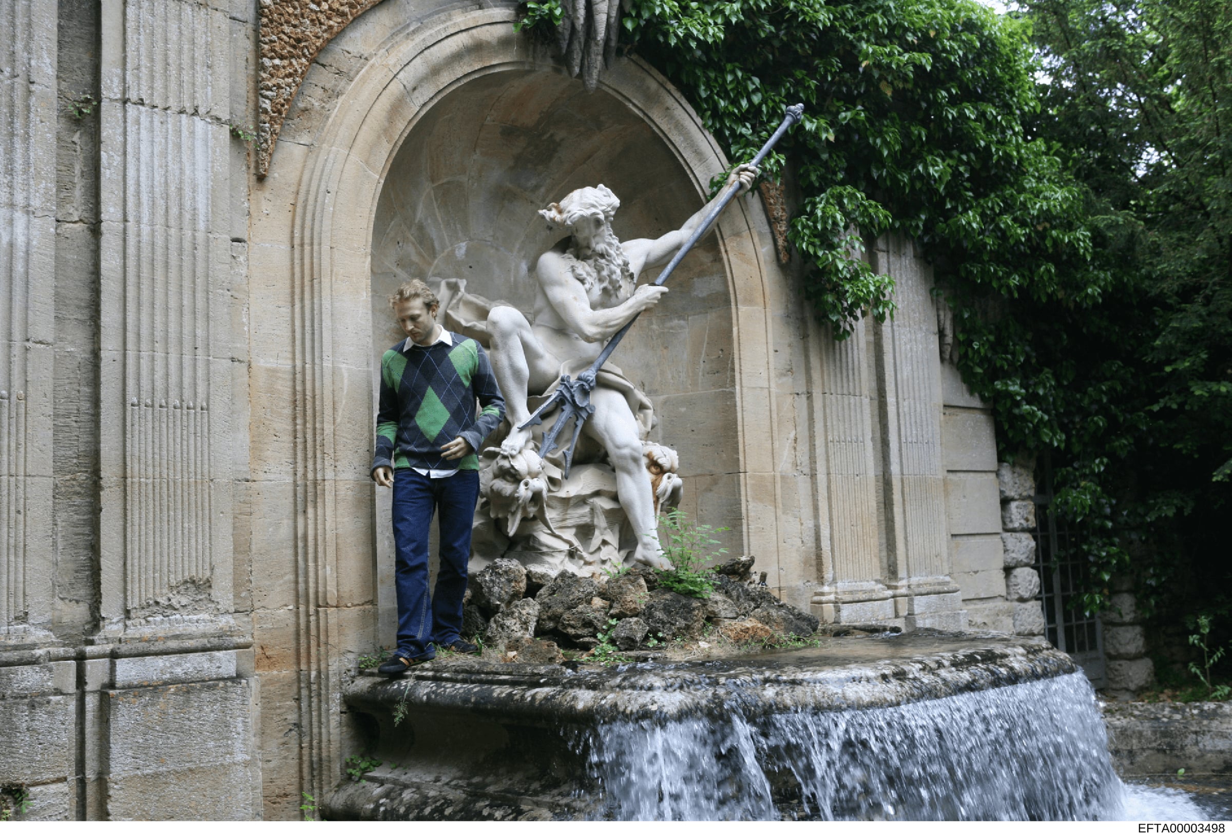 This photograph shows an unidentified male standing in front of a classical stone statue in what appears to be a European courtyard or alcove. The setting features neoclassical architecture with arched doorways, stone columns, and ivy-covered walls.