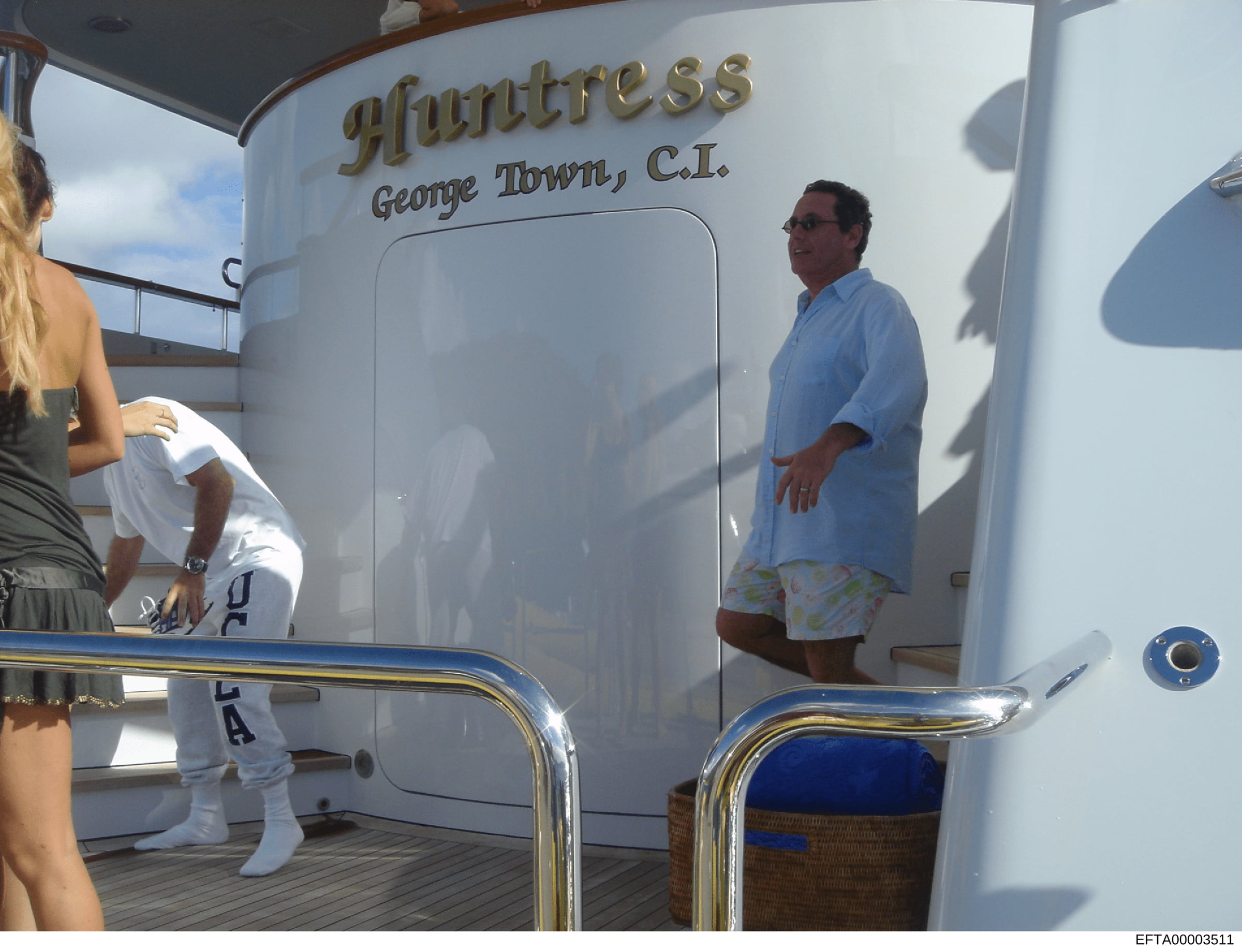 This is an evidence photograph showing a man in casual clothing standing on the deck of a yacht named 'Huntress' registered in George Town, Cayman Islands. The image appears to be from an investigation into properties and assets, showing leisure acti