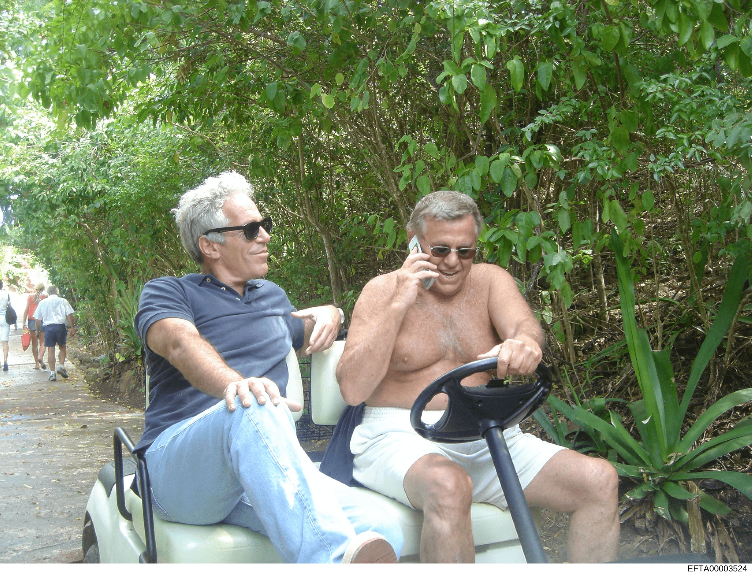 This is an evidence photograph showing two men seated on a golf cart in a lush, tree-lined tropical or subtropical location. The man on the left wears a dark blue polo shirt and sunglasses; the man on the right is shirtless, wearing white shorts and