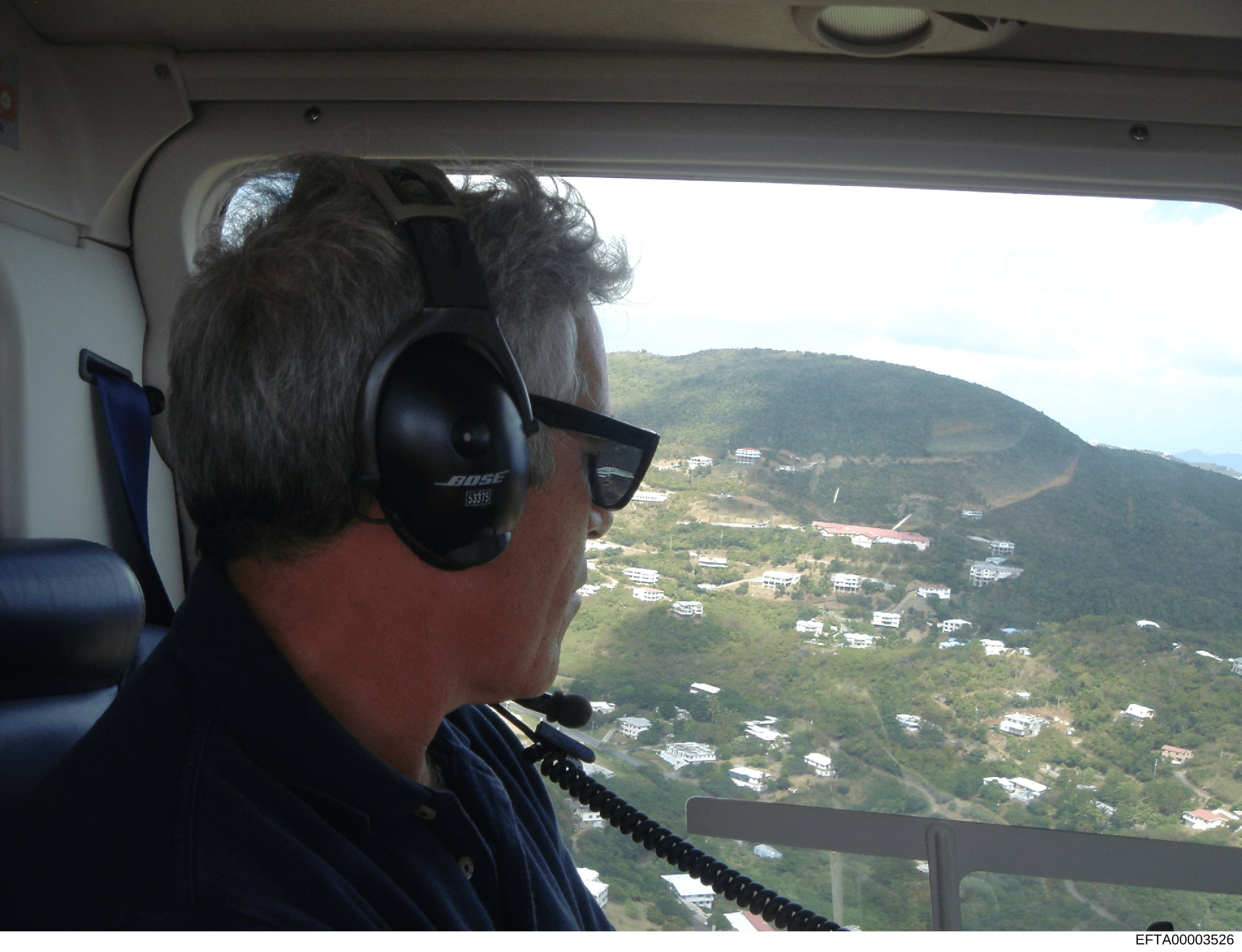 This is an aerial photograph taken from inside a helicopter, showing a person wearing headphones and sunglasses looking out at a mountainous landscape dotted with residential properties. The image appears to be documentation of aerial surveillance or