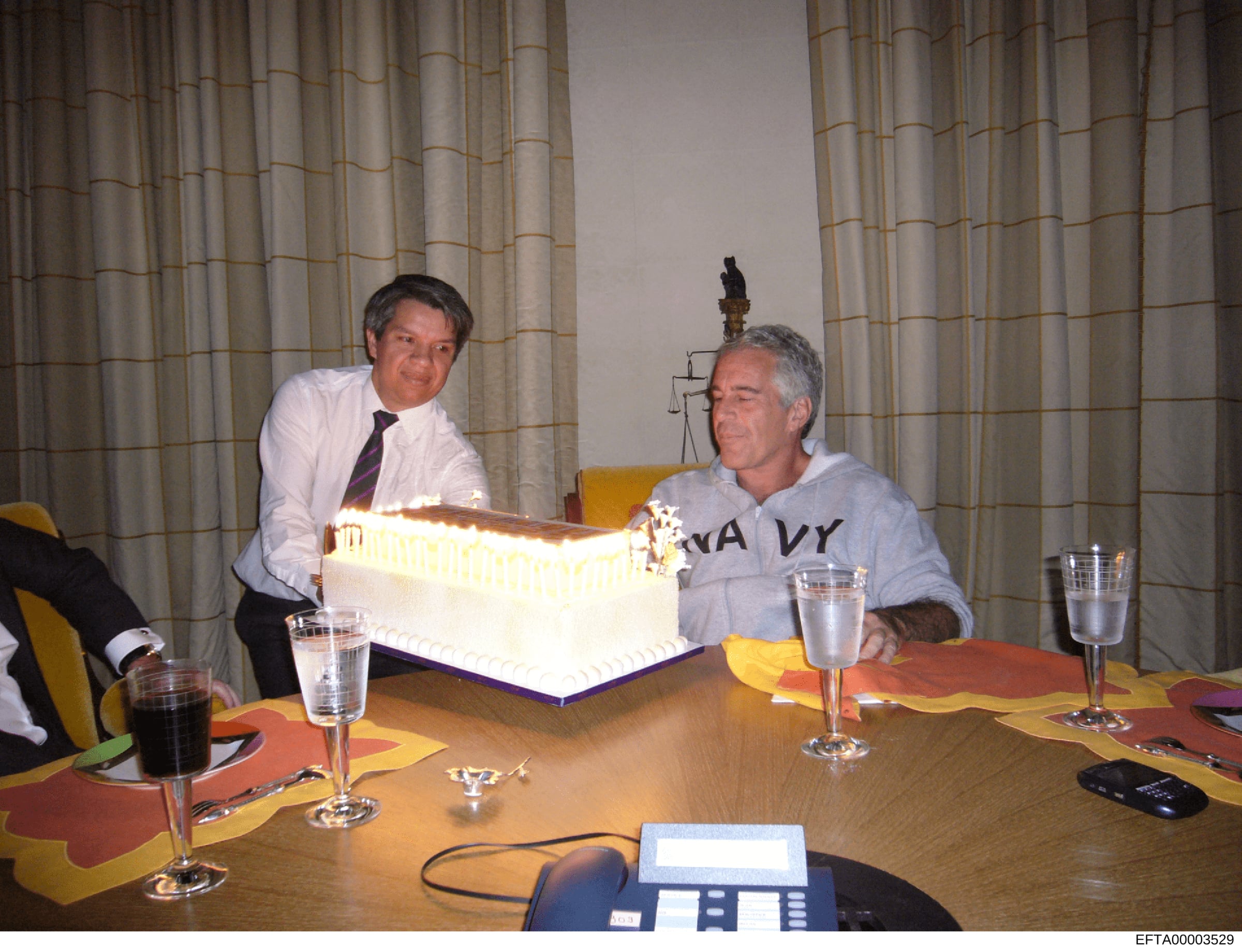 This photograph shows an indoor social or business meeting with two men seated at a wooden conference table. One man in professional attire (white shirt and tie) is on the left, while another man in casual gray clothing is on the right. A decorated w
