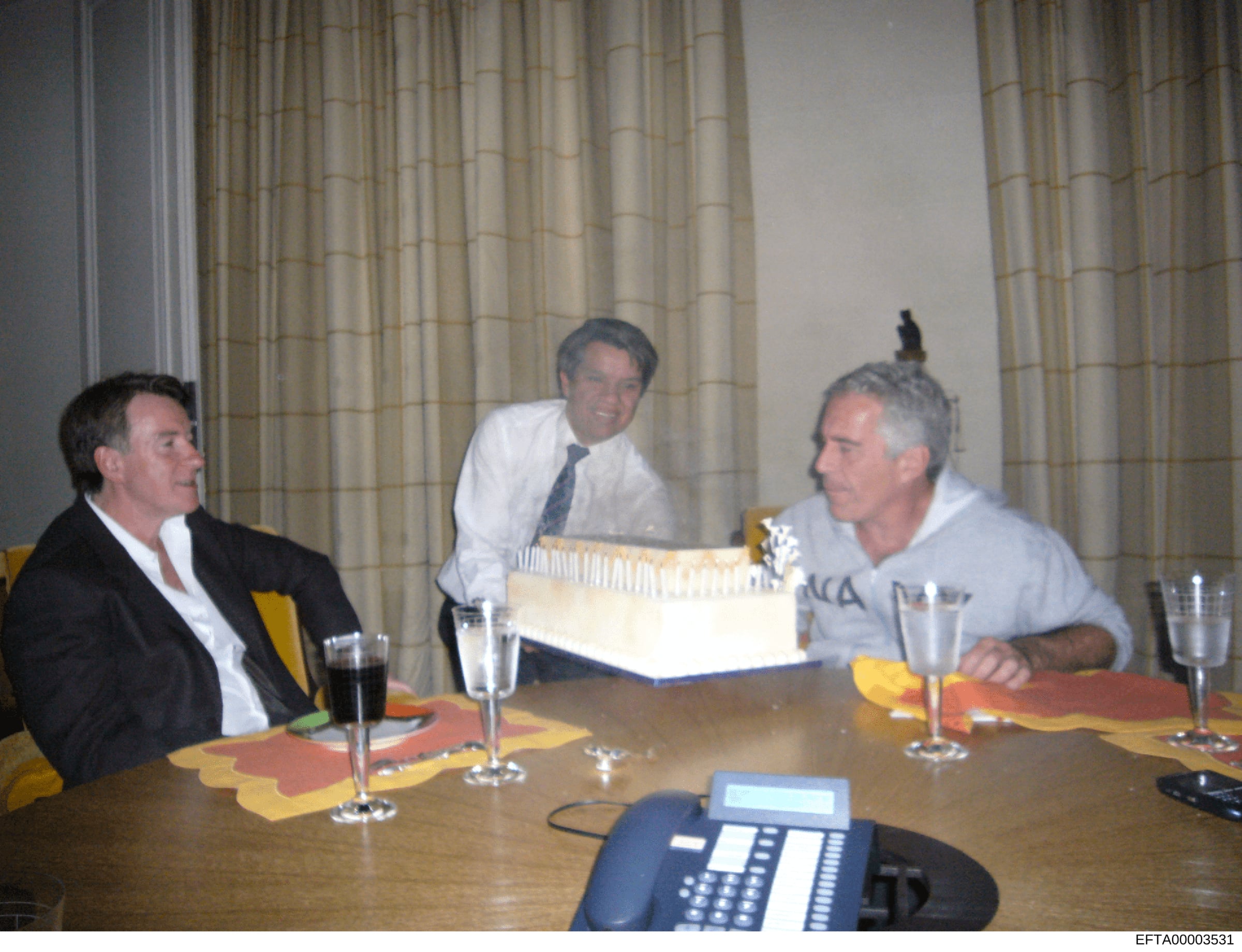 This photograph depicts three men in business attire seated at a conference table during what appears to be a formal meeting or discussion. The image shows an indoor setting with formal curtains and a cake or decorated item on the table, suggesting a