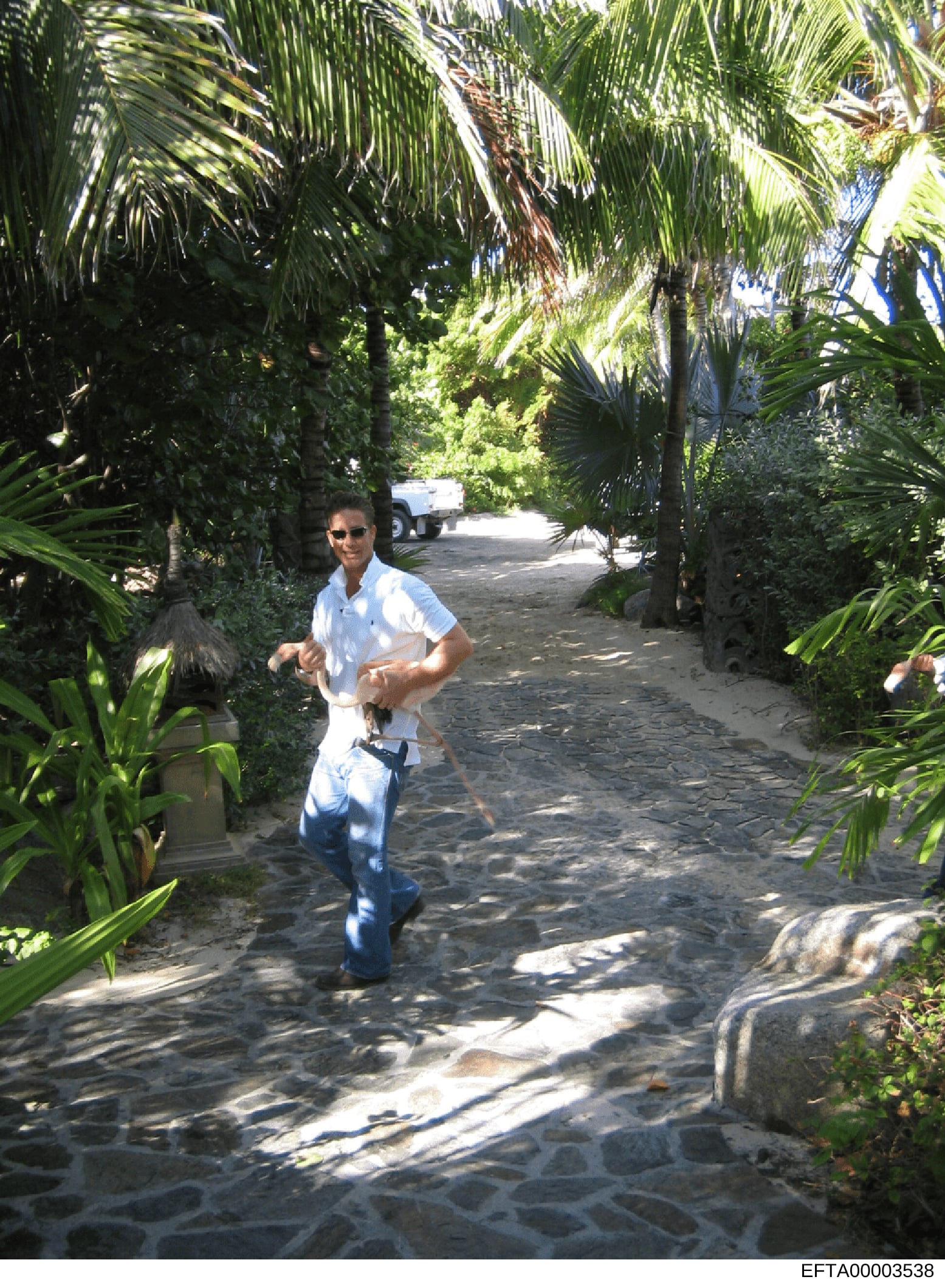 A photograph showing an unidentified man jogging on a stone driveway lined with tall palm trees and dense tropical vegetation. A vehicle is visible in the background. This appears to be evidence photo documentation of a residential property, likely o