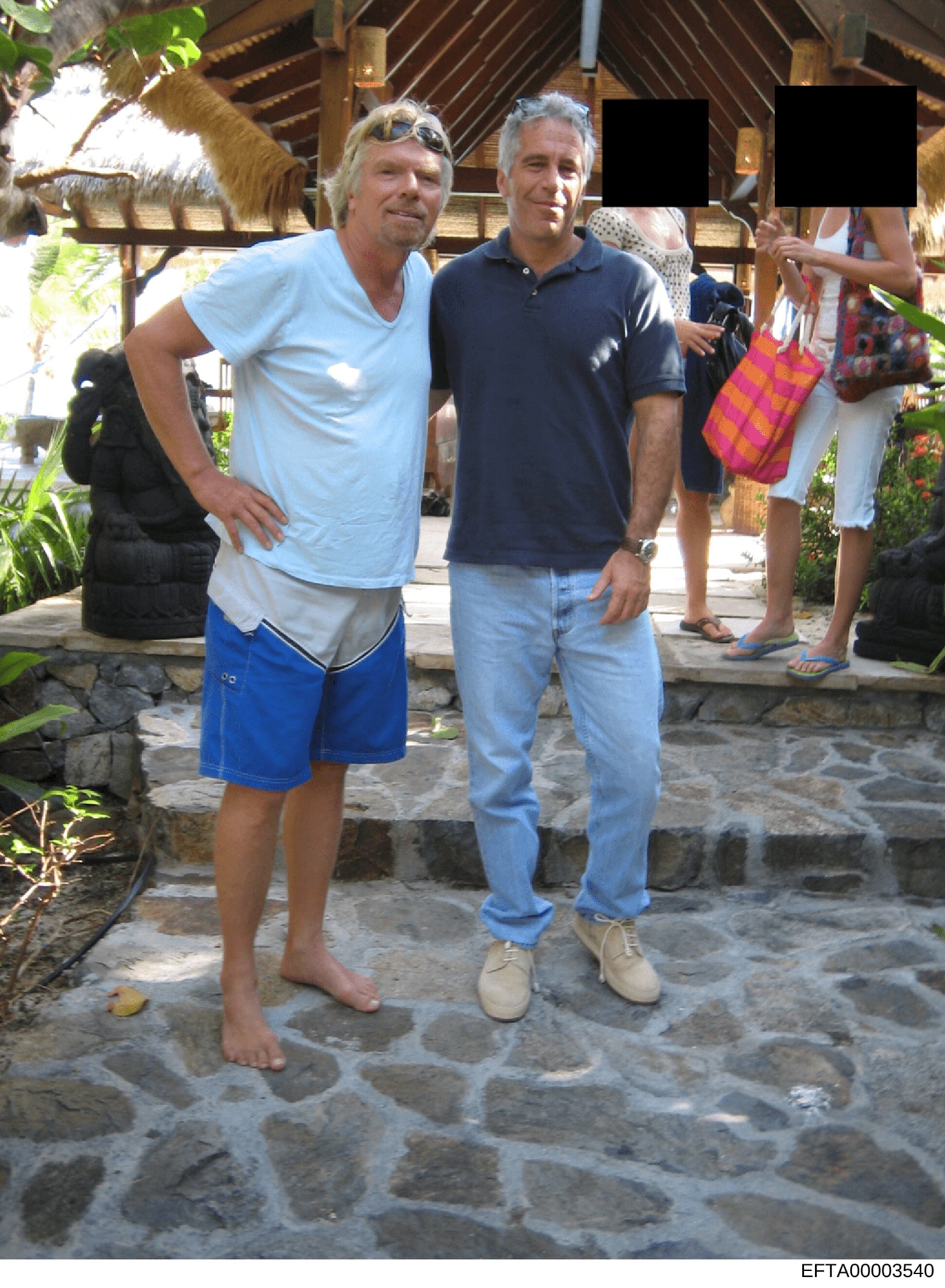 This is an evidence photograph showing two men posing together at what appears to be a tropical resort or villa with a wooden pavilion structure. The setting includes stone flooring, tropical vegetation, and other people visible in the background. Th