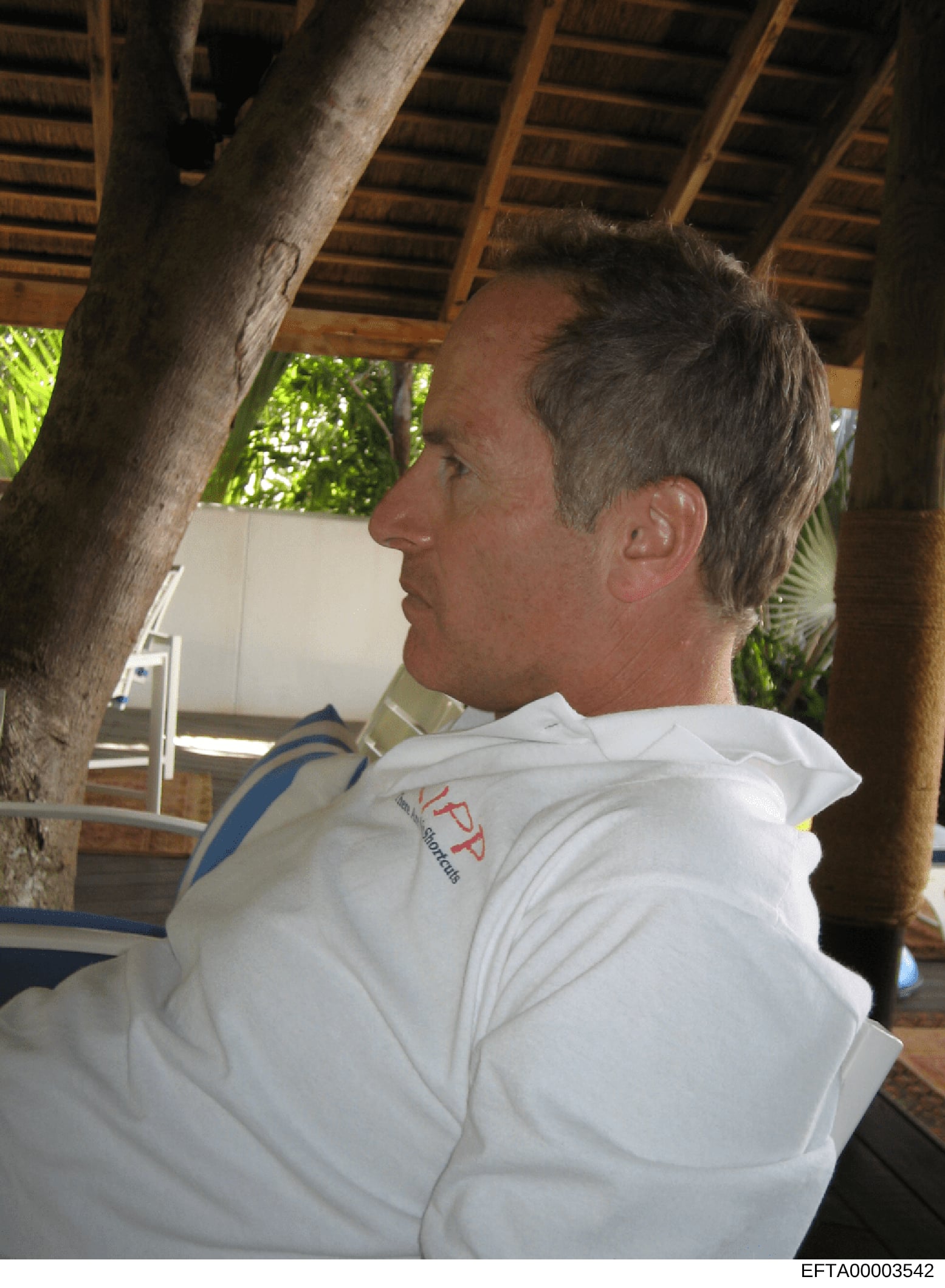 This is a photograph showing a middle-aged man wearing a white polo shirt with a red and blue logo/emblem, photographed in profile at what appears to be a covered outdoor structure or pavilion with wooden beams and tropical vegetation visible. The im