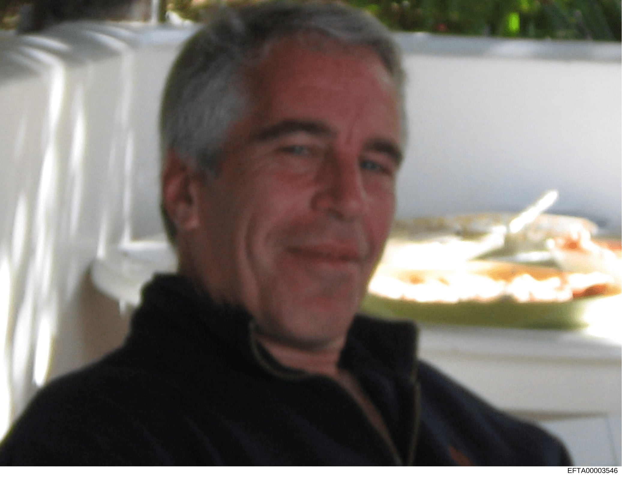 This is a photograph showing a middle-aged man with gray hair wearing dark clothing, photographed outdoors near what appears to be a catered event or gathering. A white structure is visible in the background along with plates of food. The image appea
