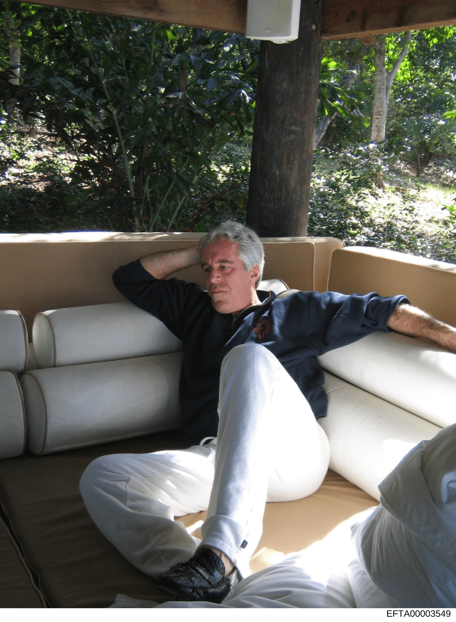 This is a photograph showing a man with gray hair relaxing on a cushioned outdoor sofa or daybed in a tropical setting. The image appears to be from an estate or property with lush vegetation visible in the background. The photograph is part of the E