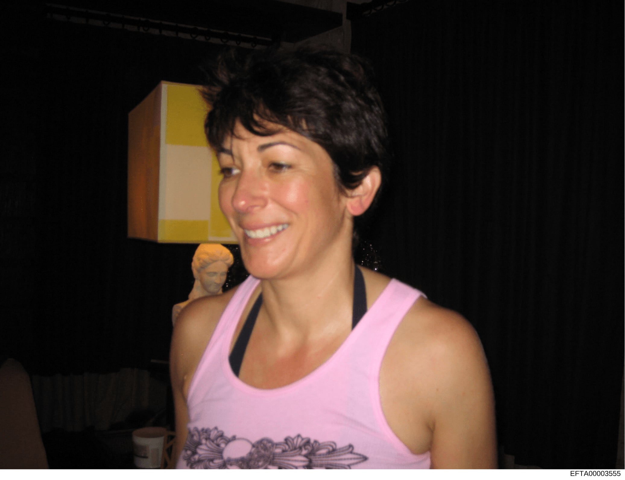 This is a photograph of a woman with short dark hair wearing a pink sleeveless top at what appears to be an indoor social gathering or event. The image is taken indoors with dark background and visible interior lighting/decor. The photo's presence in