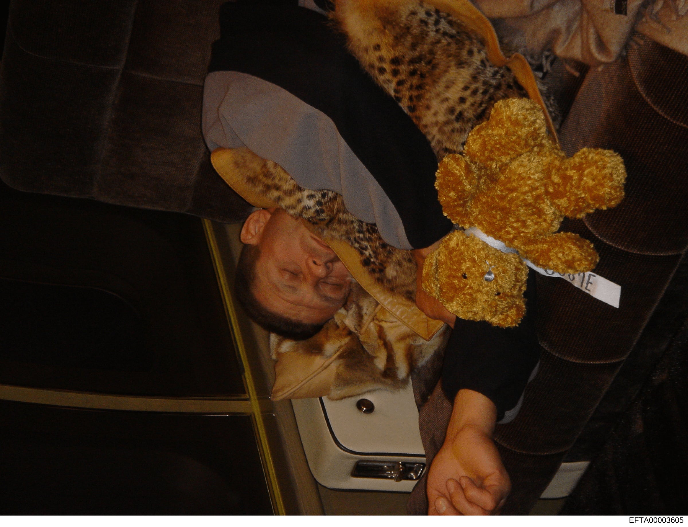This is a photograph showing a person wearing a light-colored cowboy hat with leopard print band, positioned at an angle. The person is holding or posing with a gold/yellow glittery teddy bear. The image appears to be taken indoors in low light condi