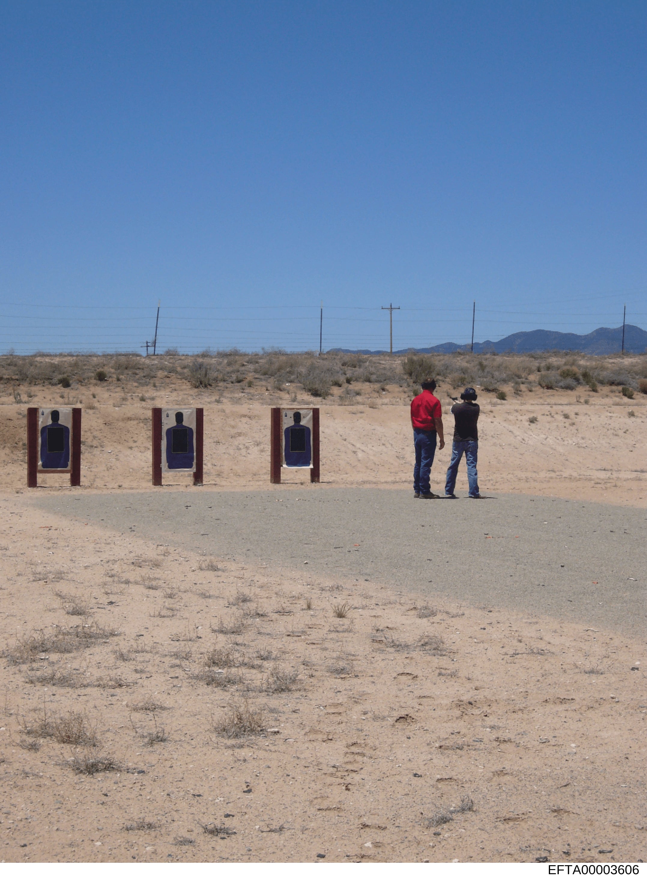This is a photograph of an outdoor shooting range in a desert landscape. The image shows three wooden target frames with blue silhouette targets, and two individuals observing from a distance. The location appears to be a remote, arid area with spars