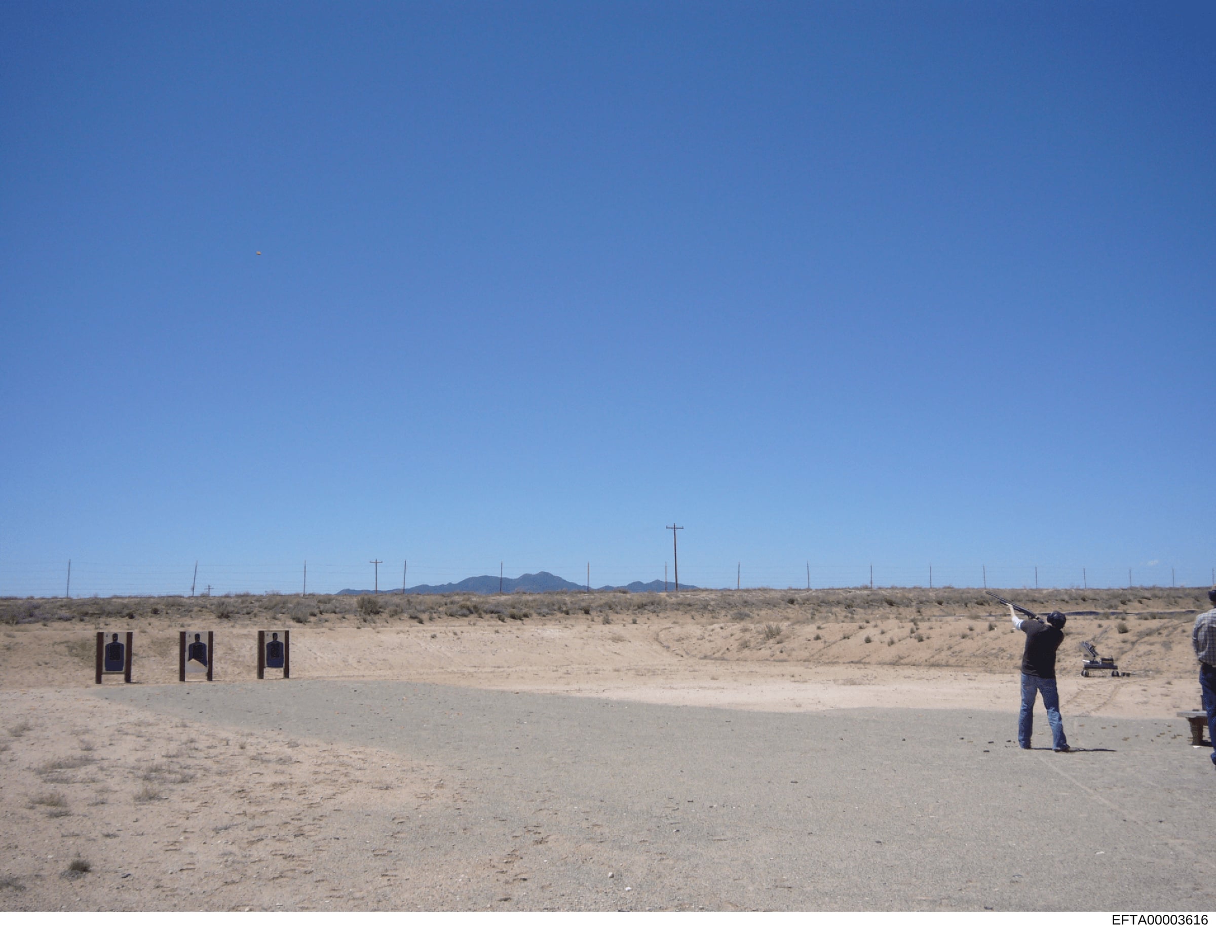 This is a photograph of an outdoor shooting range located in an arid, desert environment. The image shows three black target stands set up on a concrete pad, with a person in dark clothing positioned at a firing line. The landscape consists of sparse