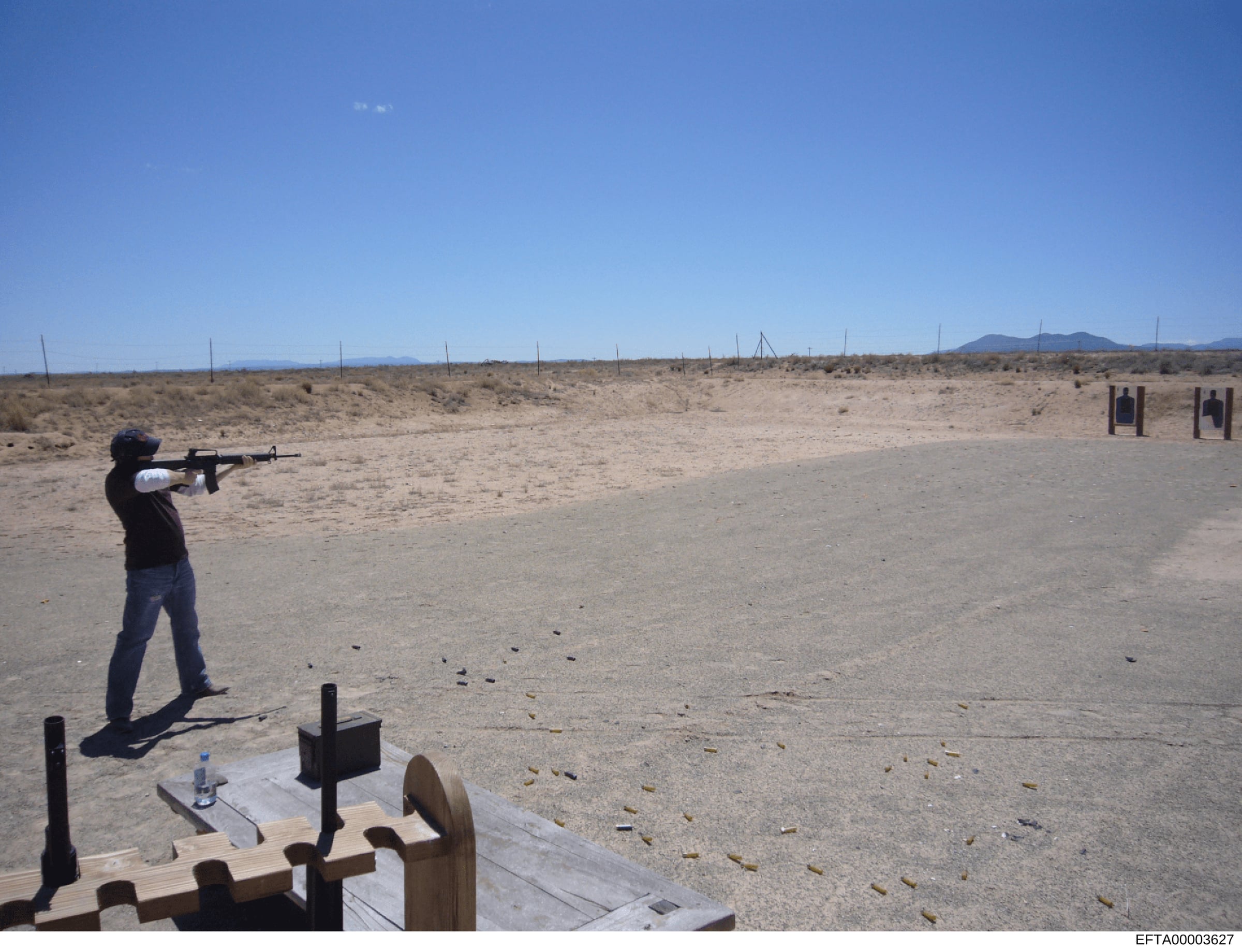 This photograph documents an individual at an outdoor desert shooting range, firing a rifle at target stations visible in the distance. The image shows spent ammunition casings on the ground and appears to be from a firearms training or practice faci