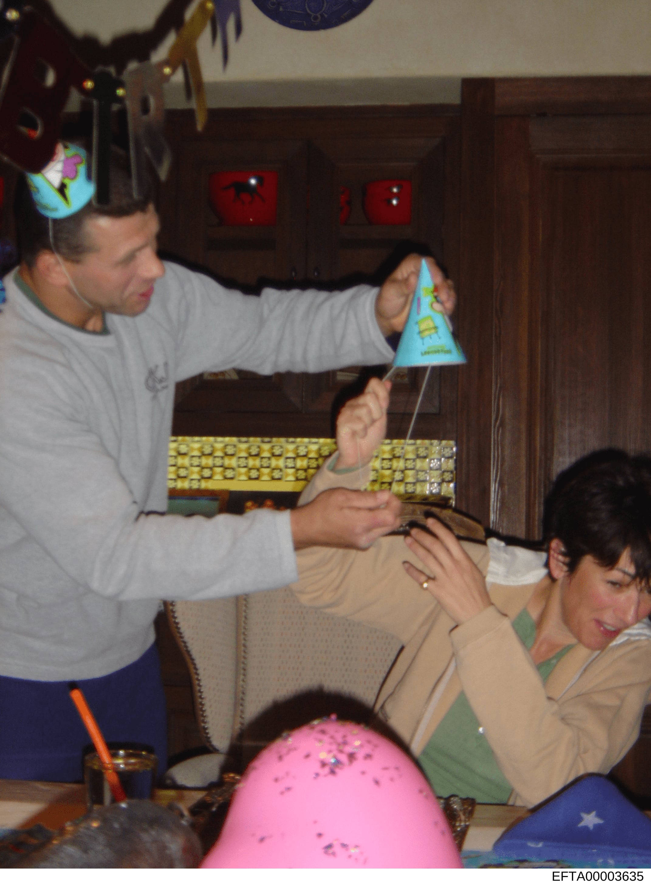This photograph shows an indoor social gathering or party, likely taking place in a residential setting. An adult male in a gray shirt is interacting with others at a table decorated with party supplies. The image captures what appears to be a casual