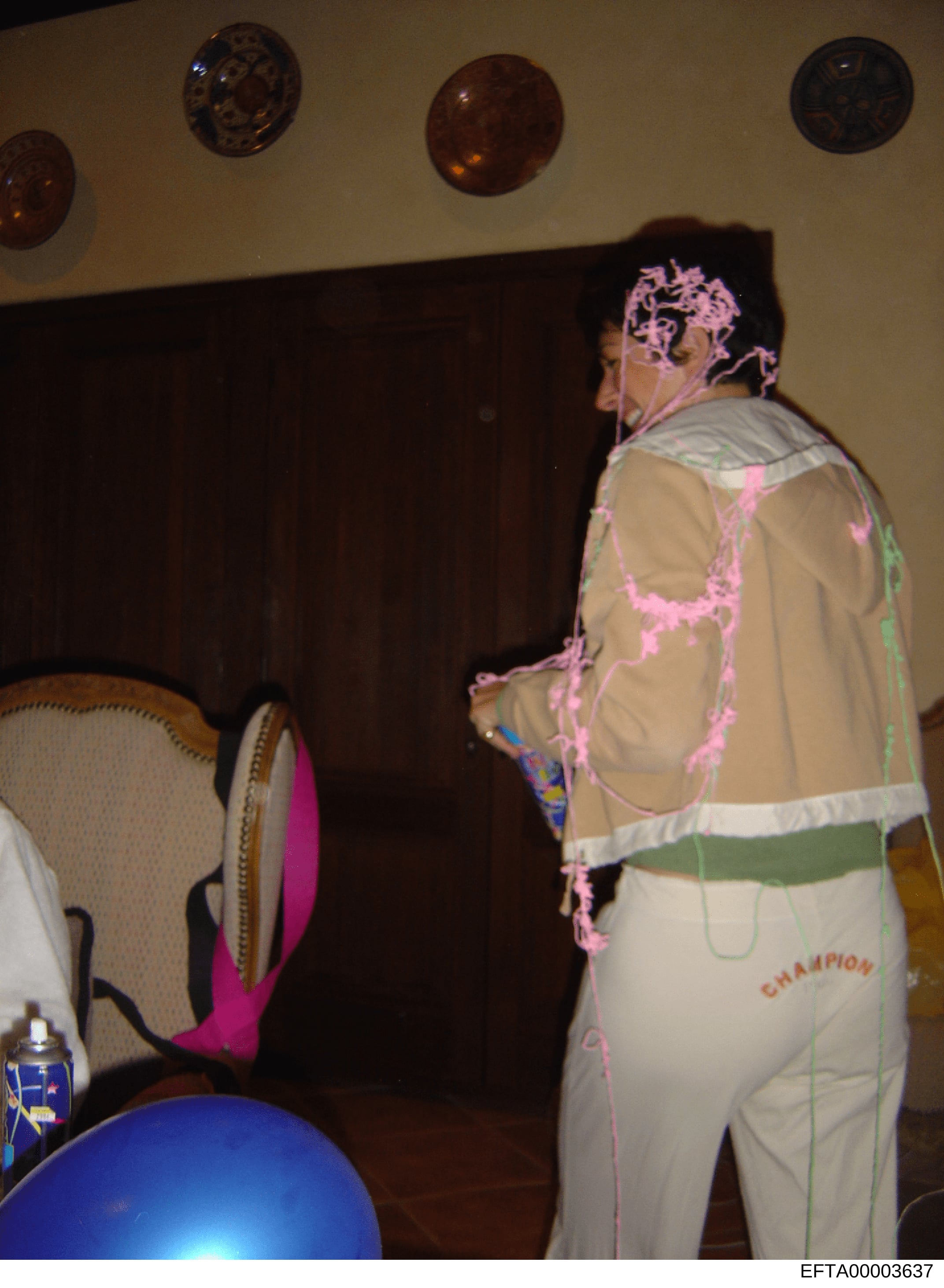 This photograph shows an interior room with a person wearing a beige/tan outfit with pink decorative elements and writing on it, along with a matching head covering. The room contains decorative circular plates on the wall, a dark curtain, and furnit