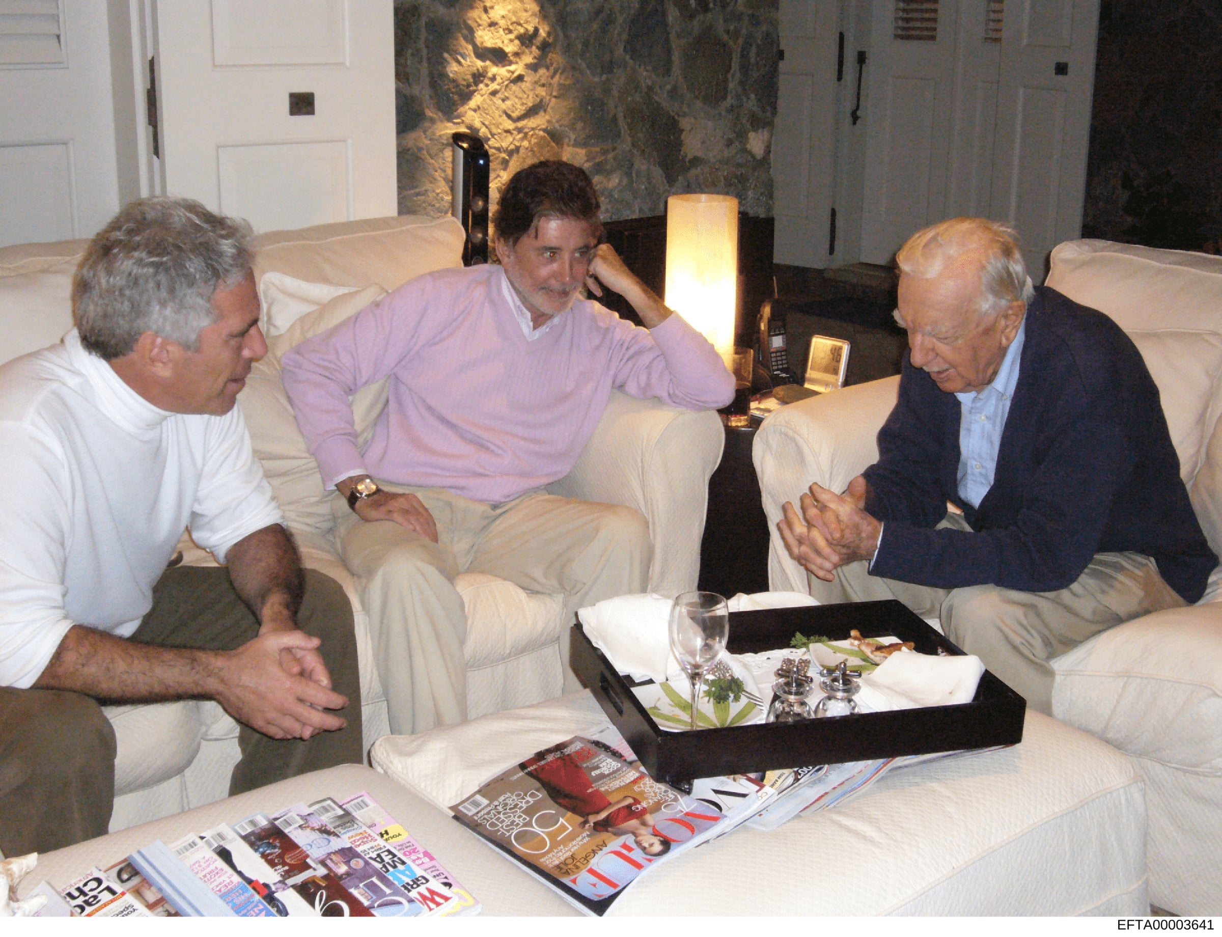 This photograph shows three men seated on cream-colored sofas in what appears to be a residential living room with contemporary furnishings and artwork. They are engaged in conversation around a black serving table with refreshments and magazines. Th
