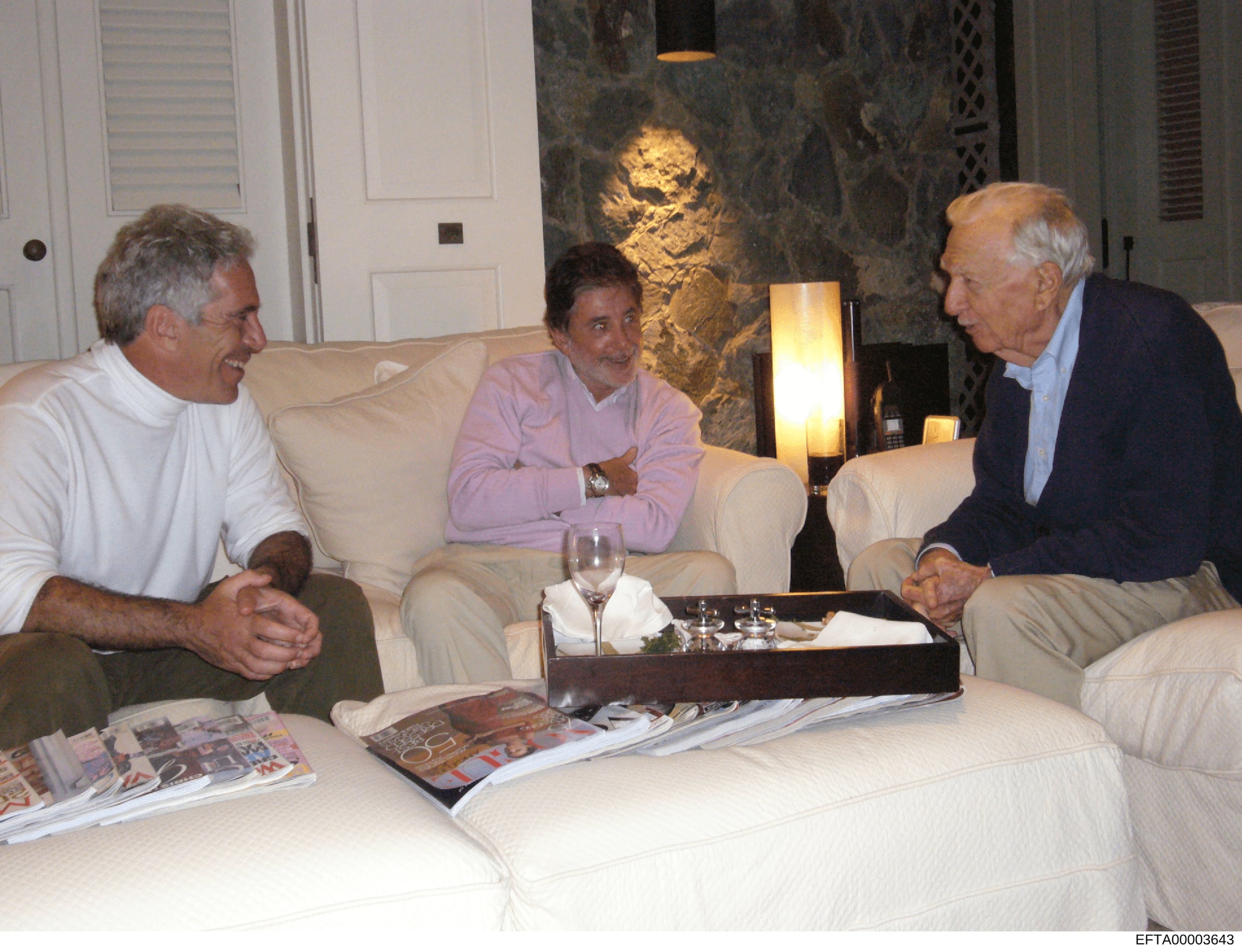 This photograph shows three men engaged in conversation in what appears to be an upscale residential living room. The setting features modern furniture, contemporary artwork, and professional lighting. The context and identities of the individuals ar