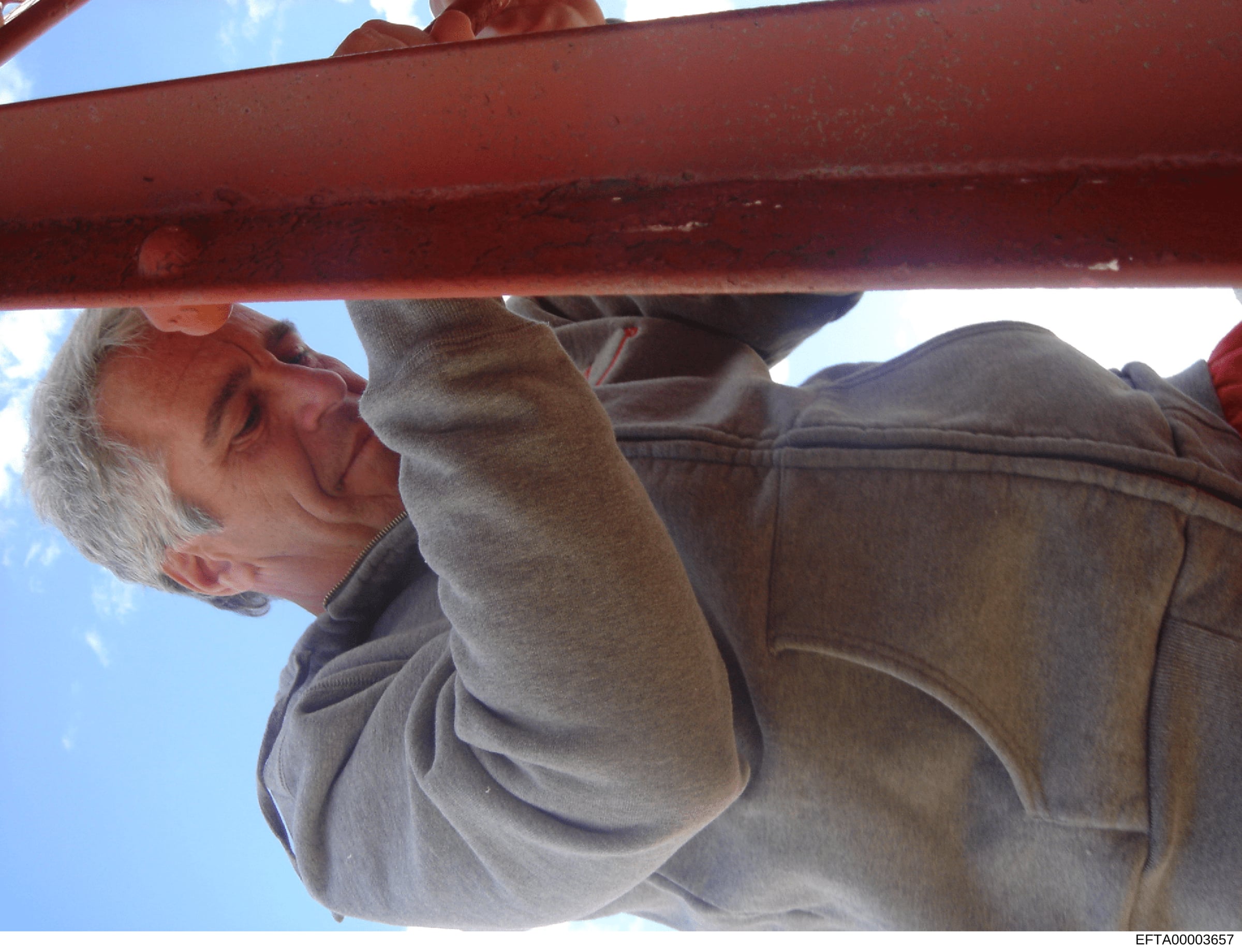 This is a candid photograph showing an older man with gray hair wearing a tan/beige jacket, positioned beneath a red metal structure or beam. The photo appears to be taken outdoors with clear sky visible in the background. The image quality and frami