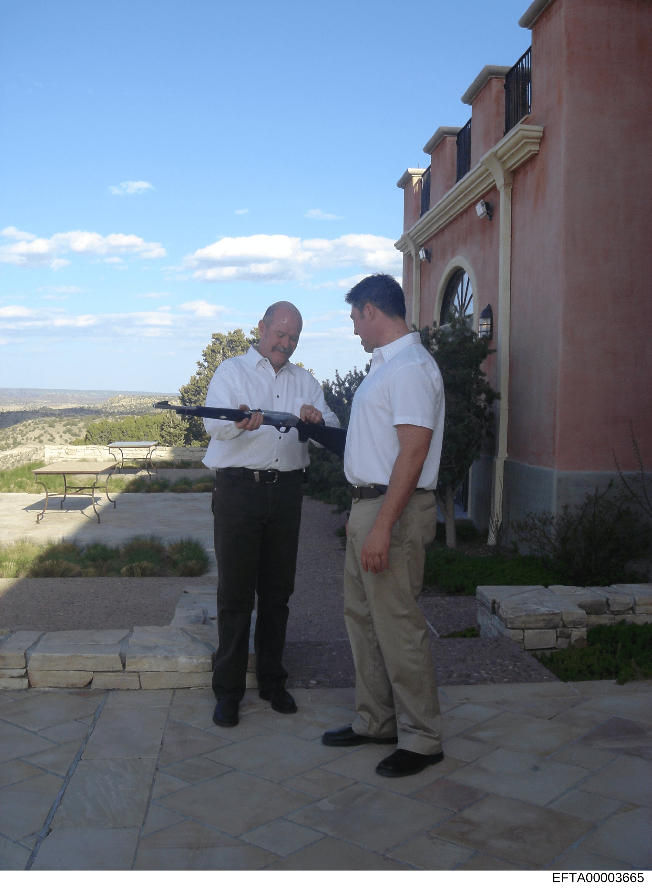 A photograph showing two men in white and light-colored shirts examining a rifle on the patio or terrace of a large residential property with southwestern architectural style. The scene appears to be taken during daytime with clear skies, overlooking
