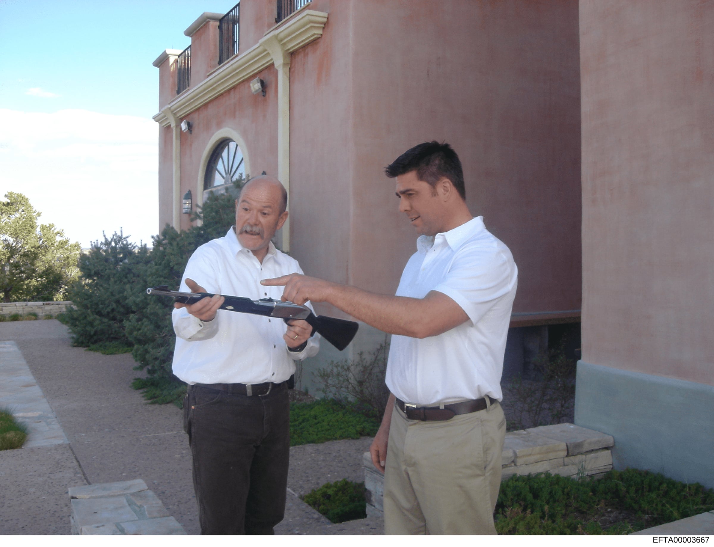 Photograph showing two men in white shirts examining a rifle outside a brick residential building. The image appears to be evidence documentation, possibly related to property inspection or weapons examination. The Bates number indicates this is part
