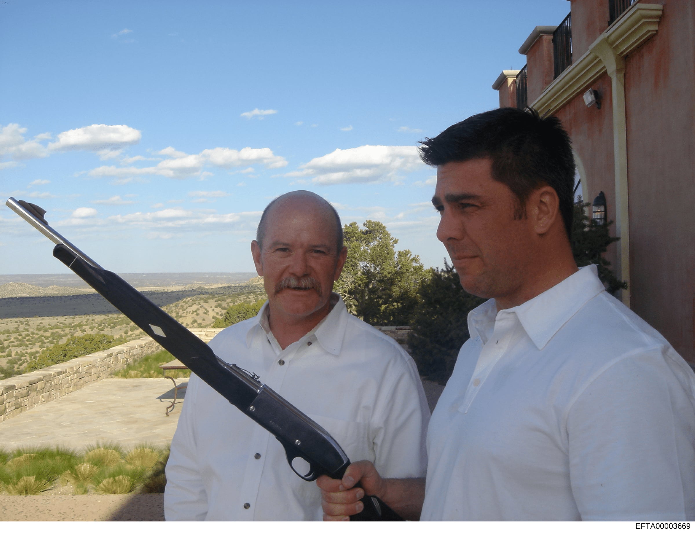 Photograph showing two unidentified men standing on a residential property terrace, with one man holding a rifle. The setting appears to be a high-elevation location with expansive views of undeveloped landscape and water in the distance. The image w