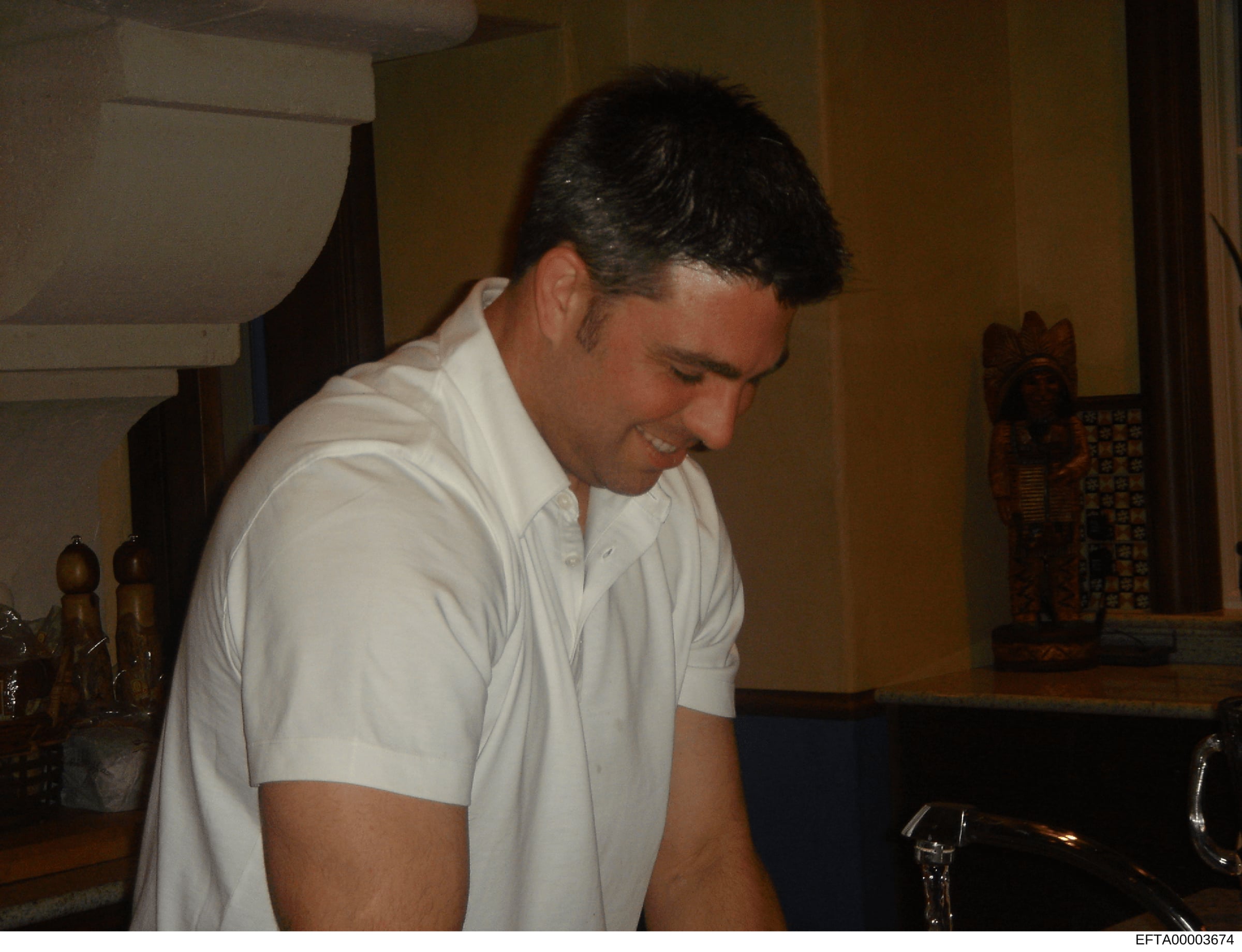 This is a candid photograph showing an unidentified man in a white short-sleeved shirt standing in what appears to be a residential interior space. The image documents an indoor setting with decorative items and furnishings visible in the background.