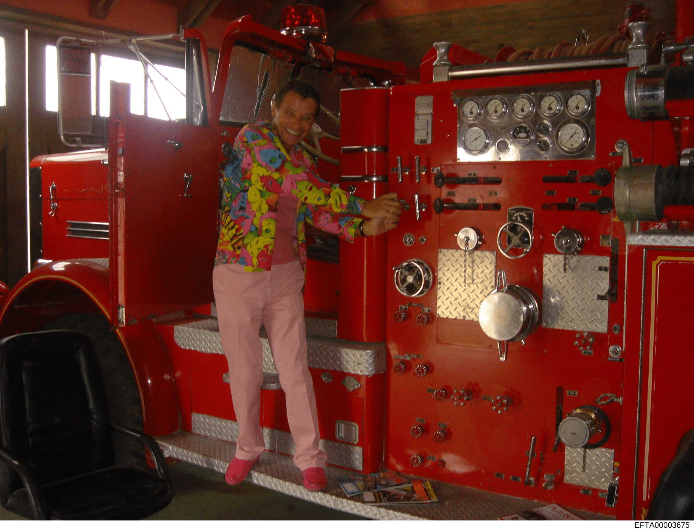 This is a casual photograph showing an unidentified person posing inside the cabin of a red fire truck. The image shows the interior equipment panel with various gauges, controls, and monitoring instruments typical of emergency response vehicles. The