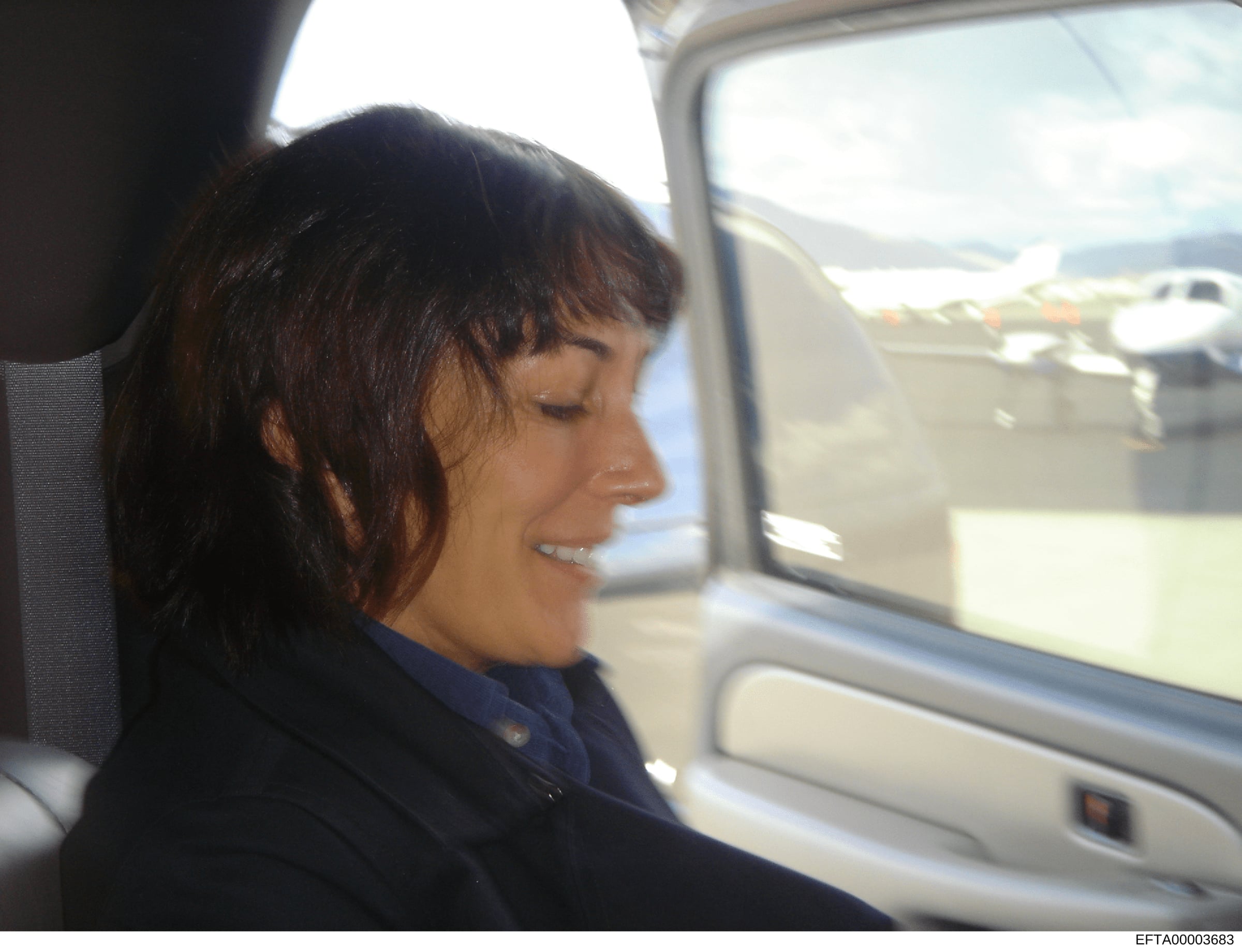 This is a candid photograph showing a woman with dark hair seated in a vehicle, photographed in profile. The setting appears to be an airport, with aircraft visible in the background through the window. The image is part of the Epstein investigation