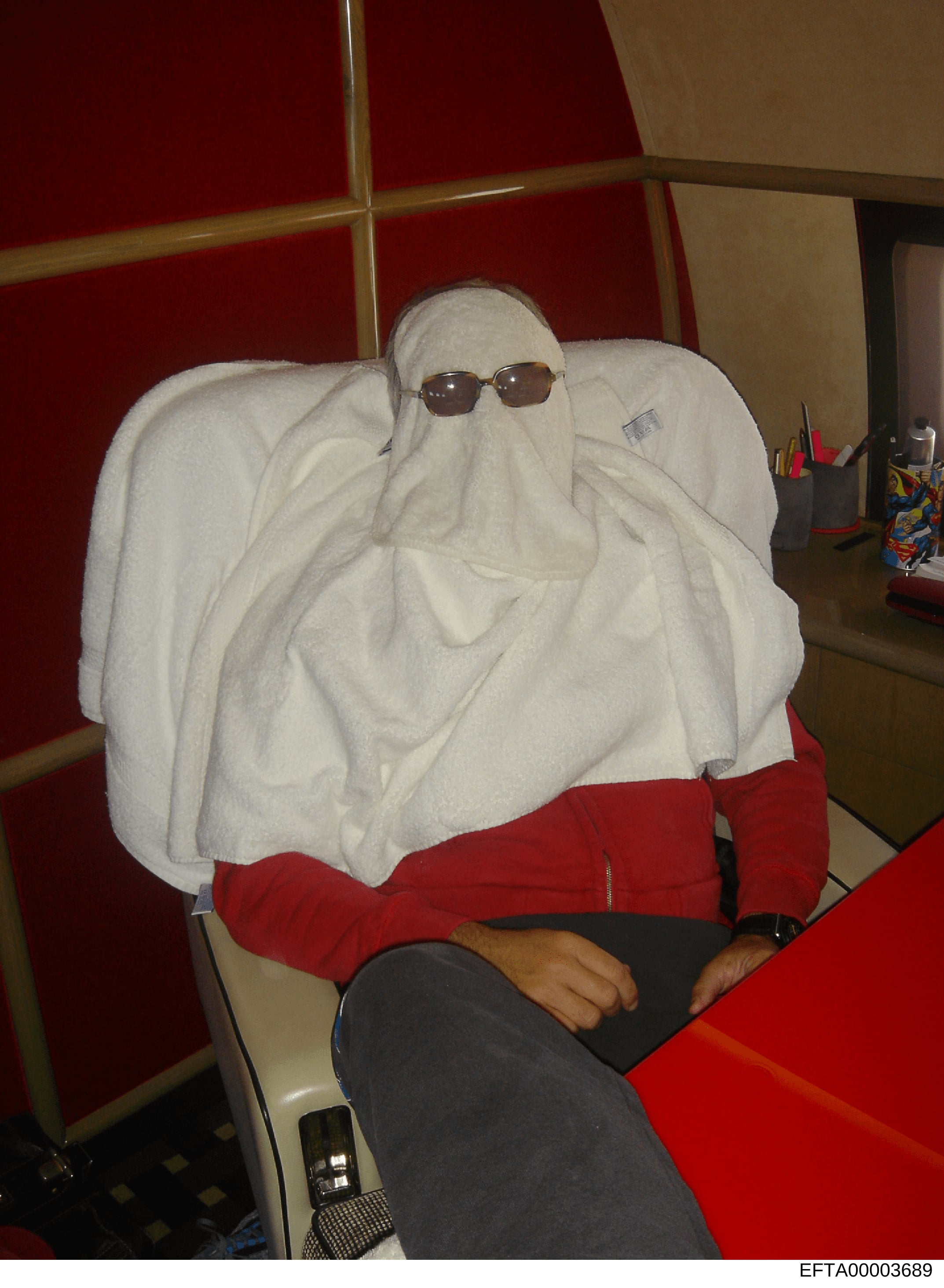 This is a photograph showing a person sitting in a chair with their head and upper body completely covered with white towels and wearing dark sunglasses, rendering their identity obscured. The image appears to be evidence documentation, possibly rela
