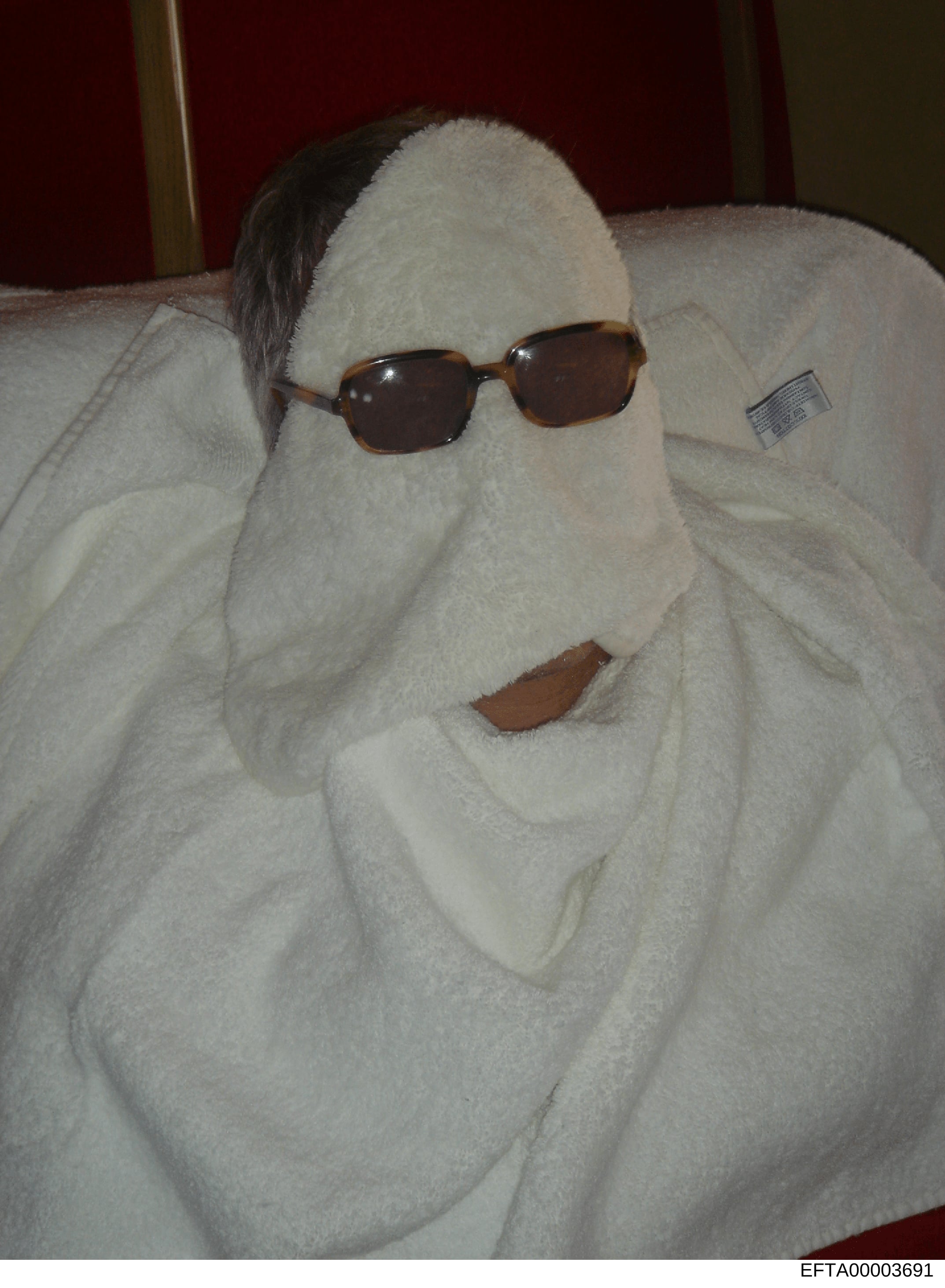 This photograph shows a white towel arranged on what appears to be a bed or furniture surface, with a pair of brown-framed sunglasses positioned on top and what appears to be a brown leather item (possibly a wallet or small accessory) visible. The im