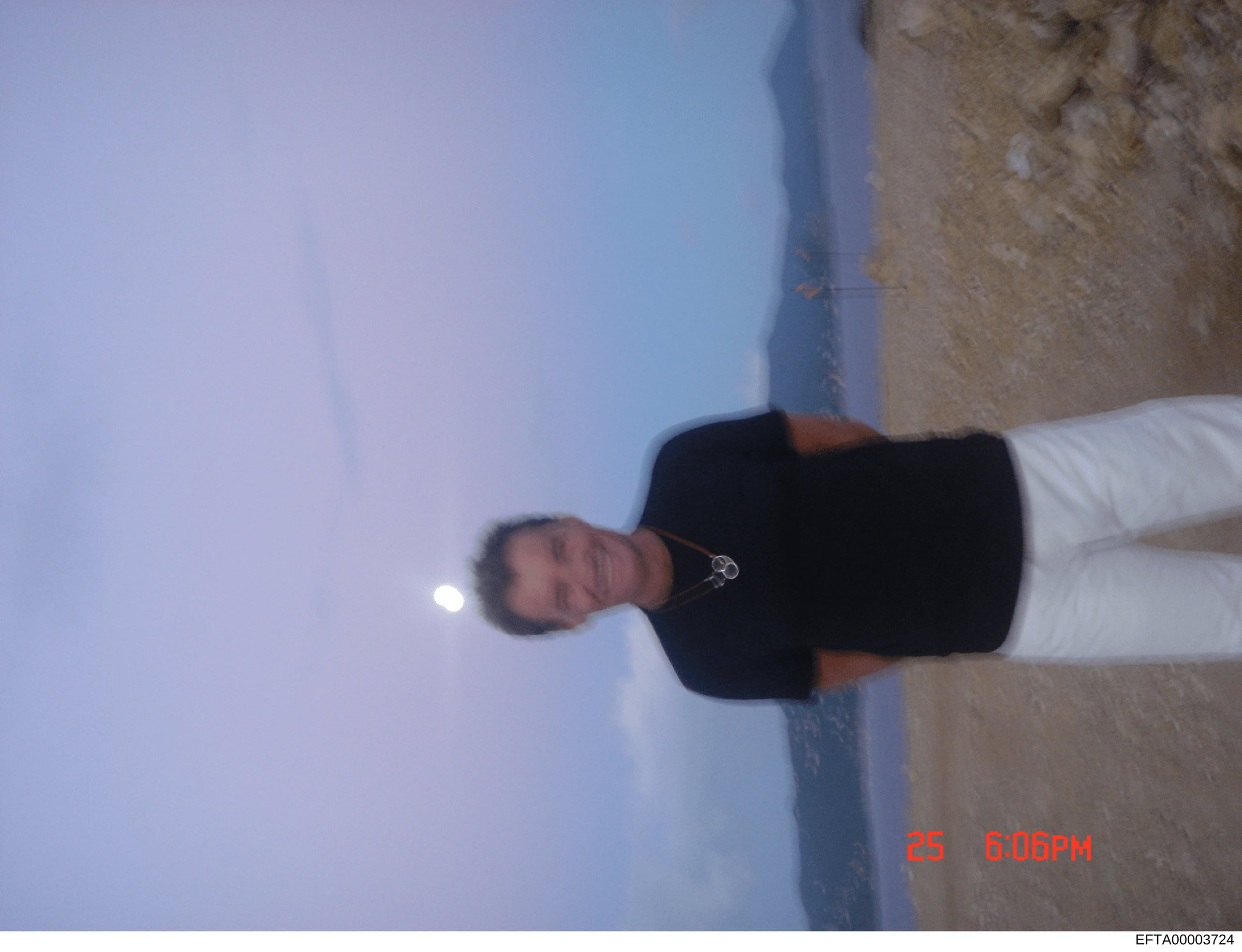 This is a candid photograph showing a person standing outdoors against a clear blue sky and stone/concrete wall background. The image is rotated 90 degrees and dated 25 June at 6:06 PM based on the timestamp visible in the lower right corner. The pho