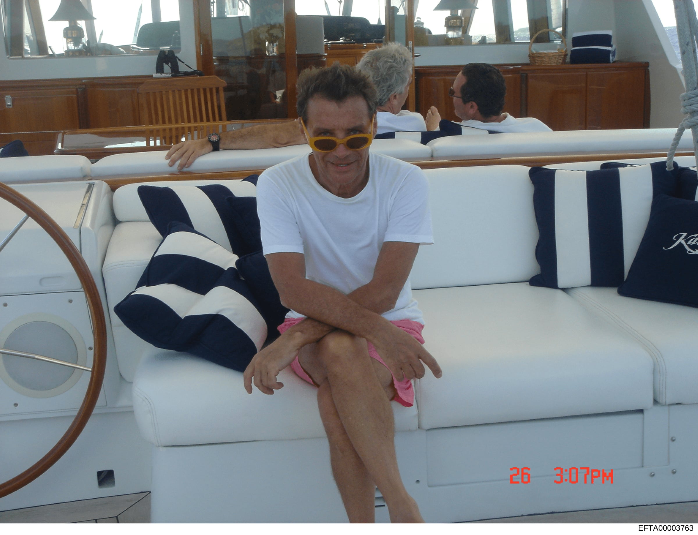A casual photograph taken on a yacht showing a man in a white t-shirt and pink shorts sitting on white upholstered seating. The photo was taken on July 26 at 3:07 PM. Two other individuals are visible in the background near the yacht's cabin. This im