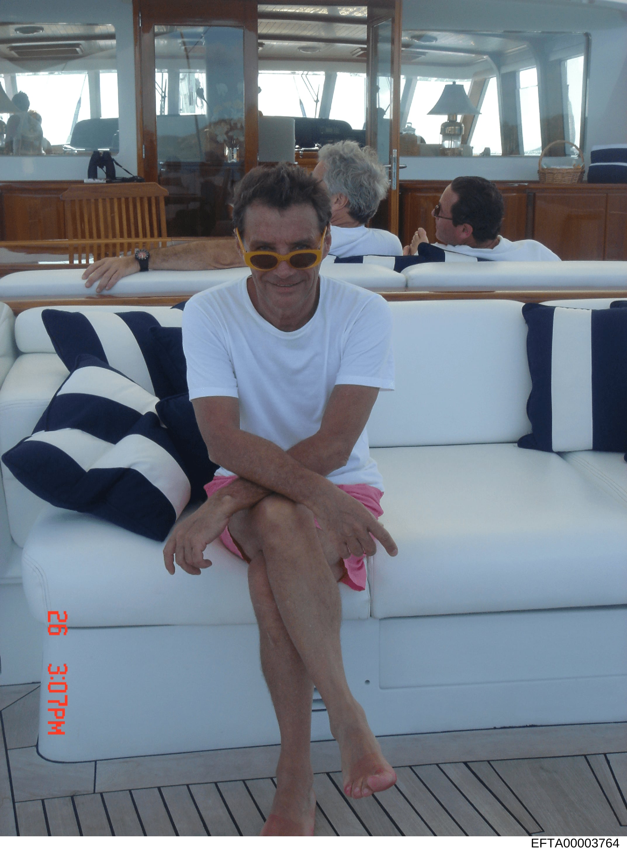 This is a photograph taken aboard a yacht showing a person in casual attire (white t-shirt, pink shorts, yellow sunglasses) seated on white upholstered seating on the deck. Two additional passengers are visible in the background. The image appears to