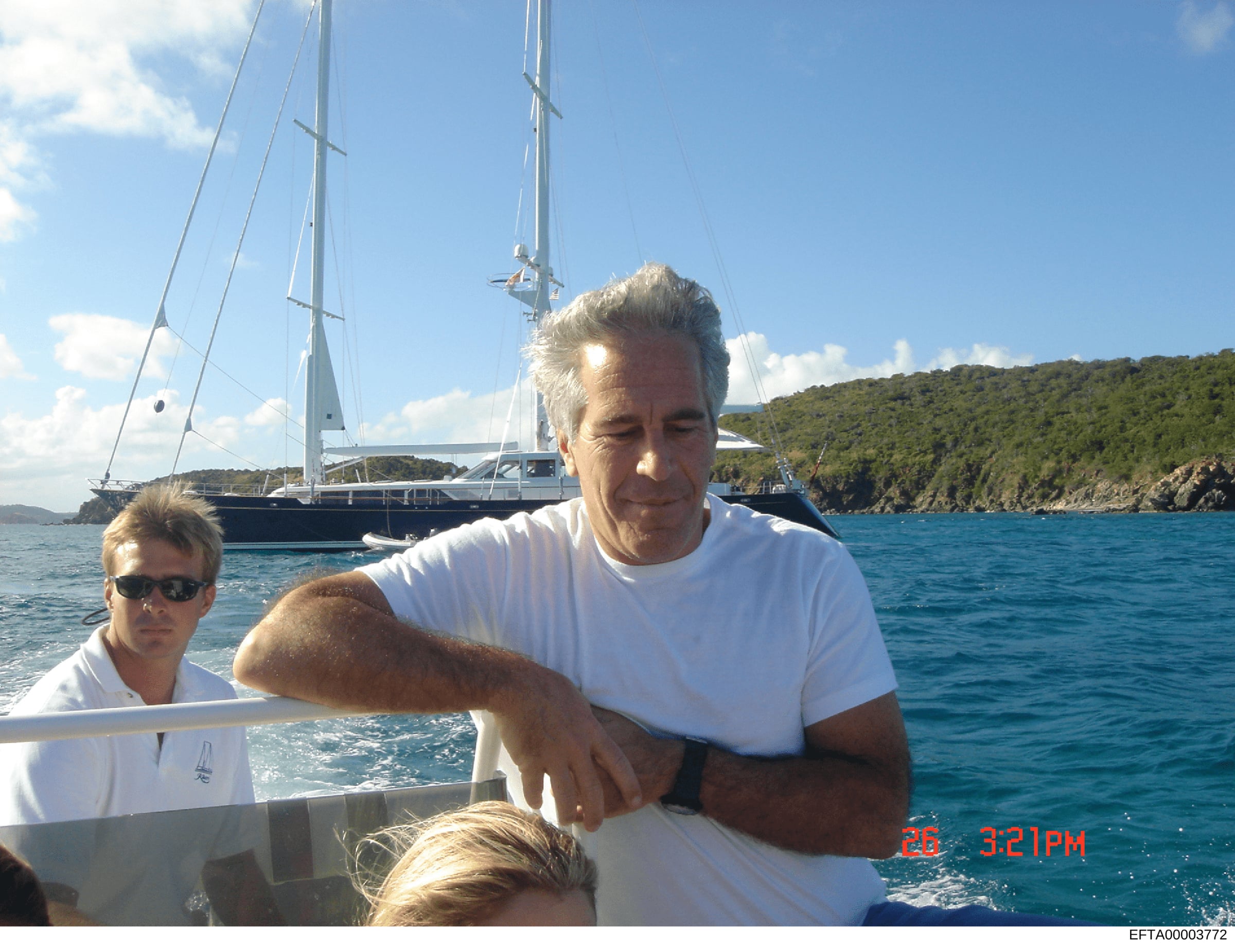 This is a personal photograph showing an older man with gray hair wearing a white t-shirt on a boat, with a large luxury sailing yacht visible in the background. A second person wearing sunglasses is partially visible to the left. The photo appears t