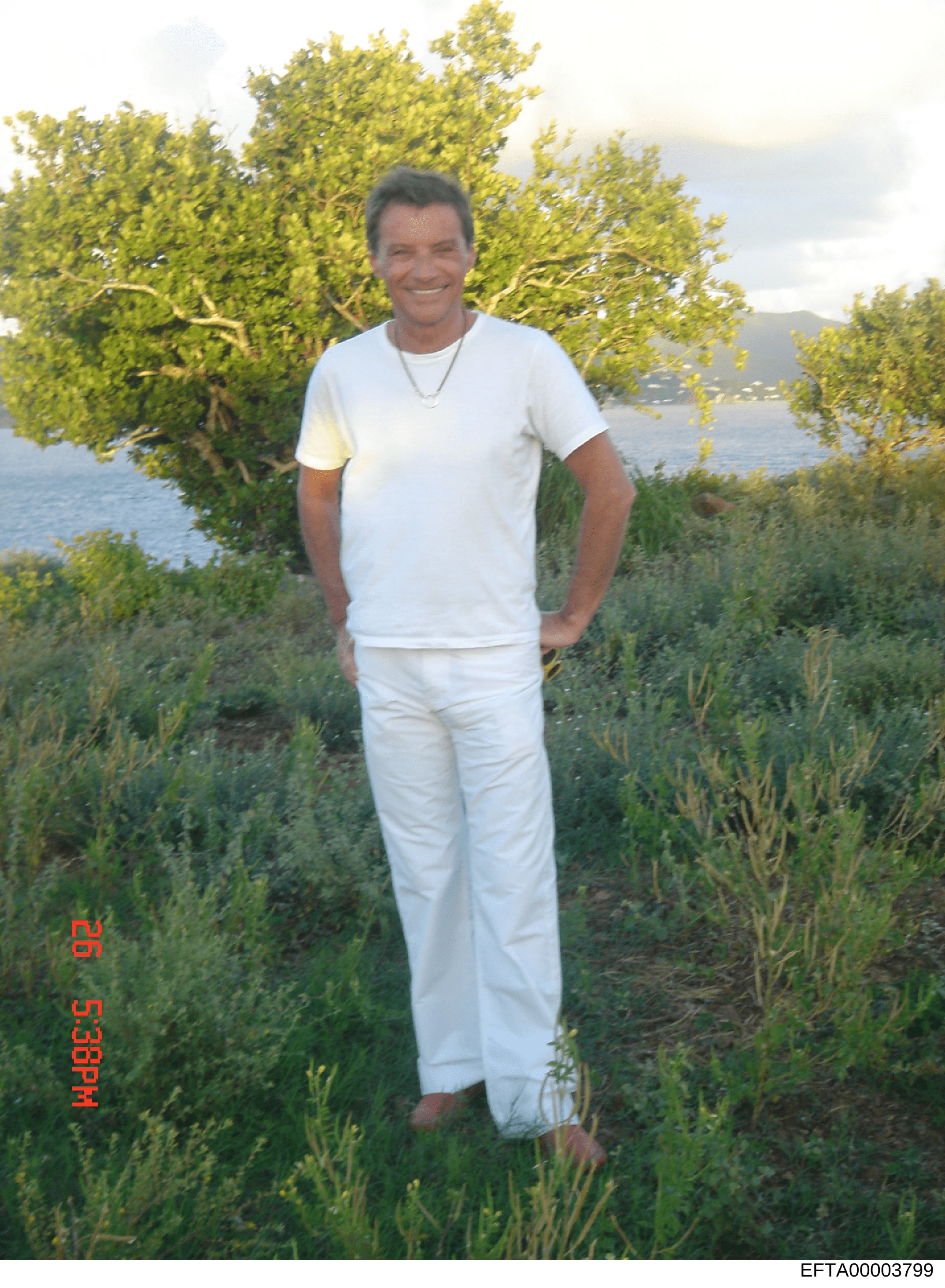 This is a casual personal photograph showing a middle-aged male wearing all-white clothing standing in a landscaped garden area overlooking a body of water. The image appears to be from the mid-1990s based on photo quality and styling. The photograph