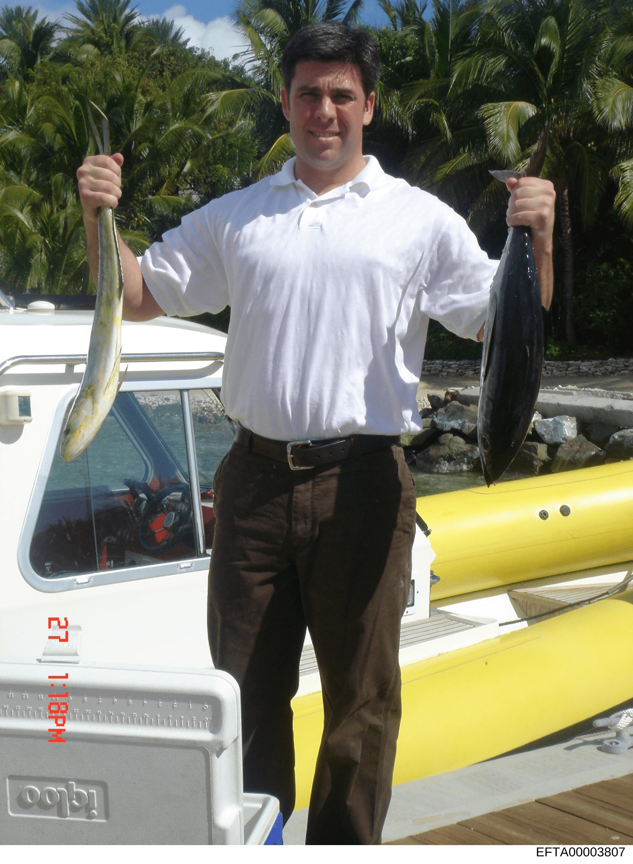 This photograph shows a man at a waterfront location displaying two caught fish while standing next to a white and yellow boat. The image appears to be from investigation files and may serve to document an individual's activities or establish presenc