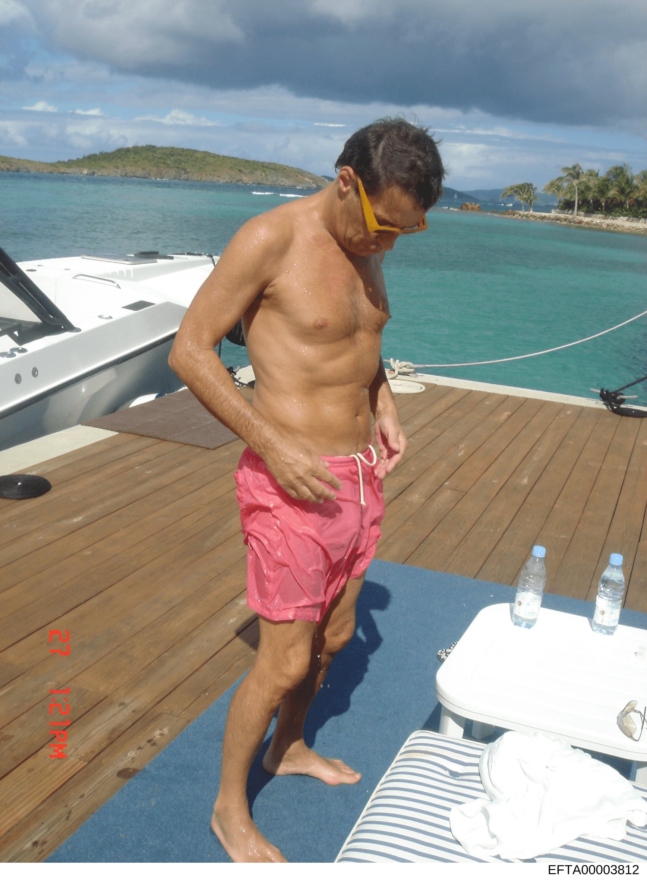 This is a personal photograph showing a shirtless man standing on the teak deck of a luxury yacht in clear tropical waters. The image appears to be casual vacation or leisure documentation, with a scenic island coastline and turquoise water visible i