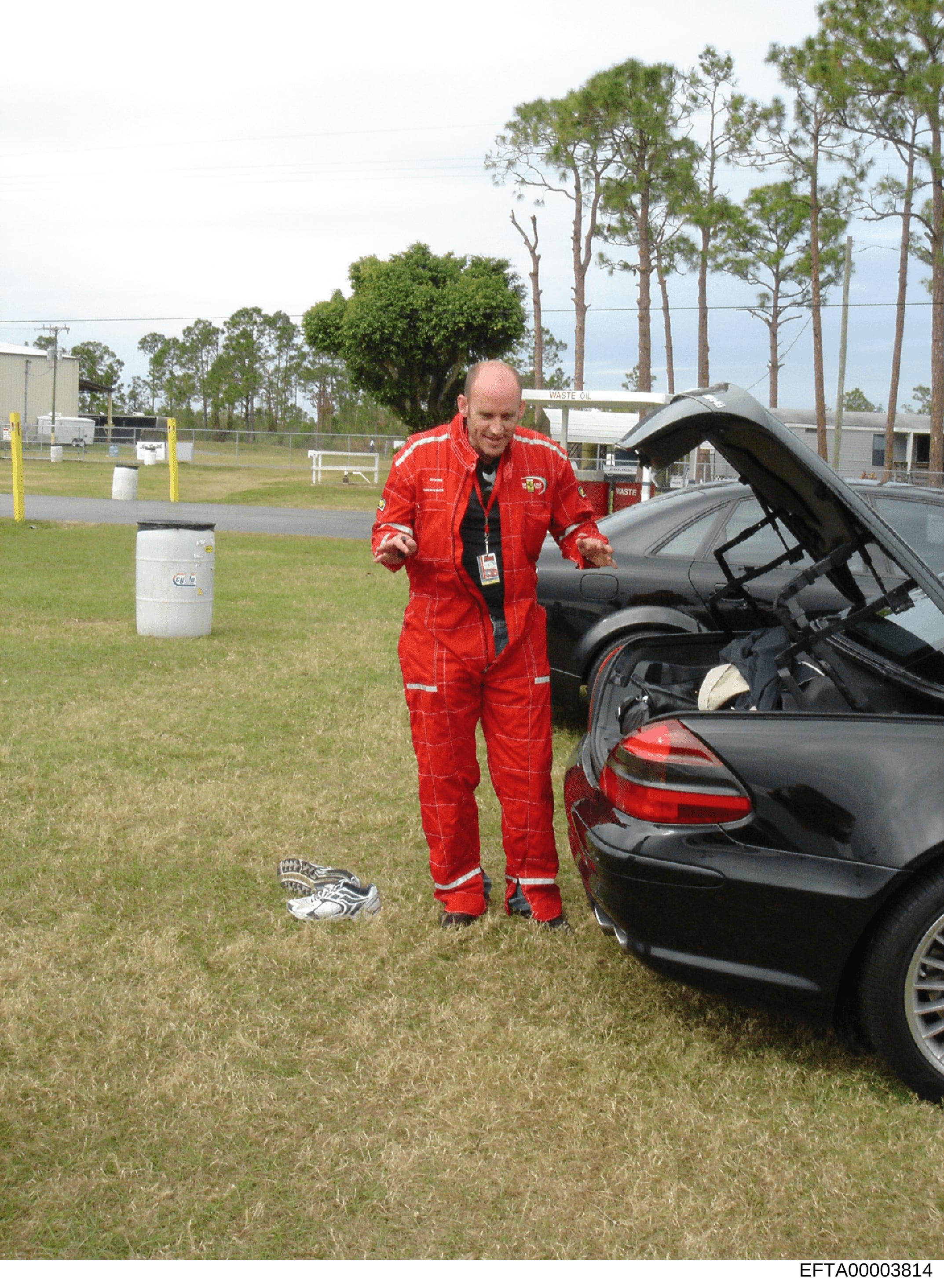 This photograph shows a man in full red firefighter protective gear and an ID badge standing next to a black sedan with its hood open at what appears to be a training or demonstration facility. The image includes industrial storage tanks, grass field
