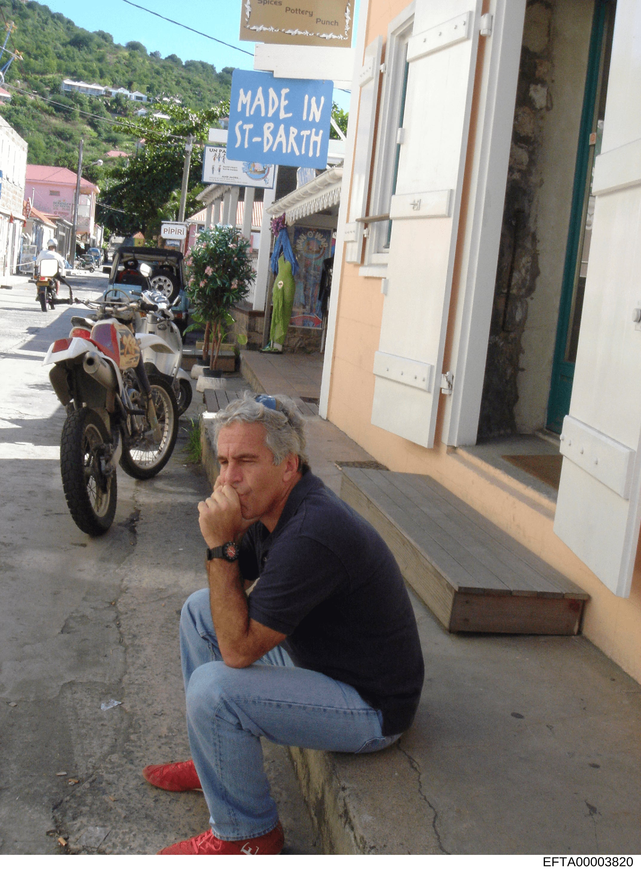 This photograph shows Jeffrey Epstein crouching on a street in Saint Barthélemy (St. Barth), a Caribbean island location. The image captures him in a casual pose wearing a navy polo shirt and jeans on what appears to be a commercial street with local