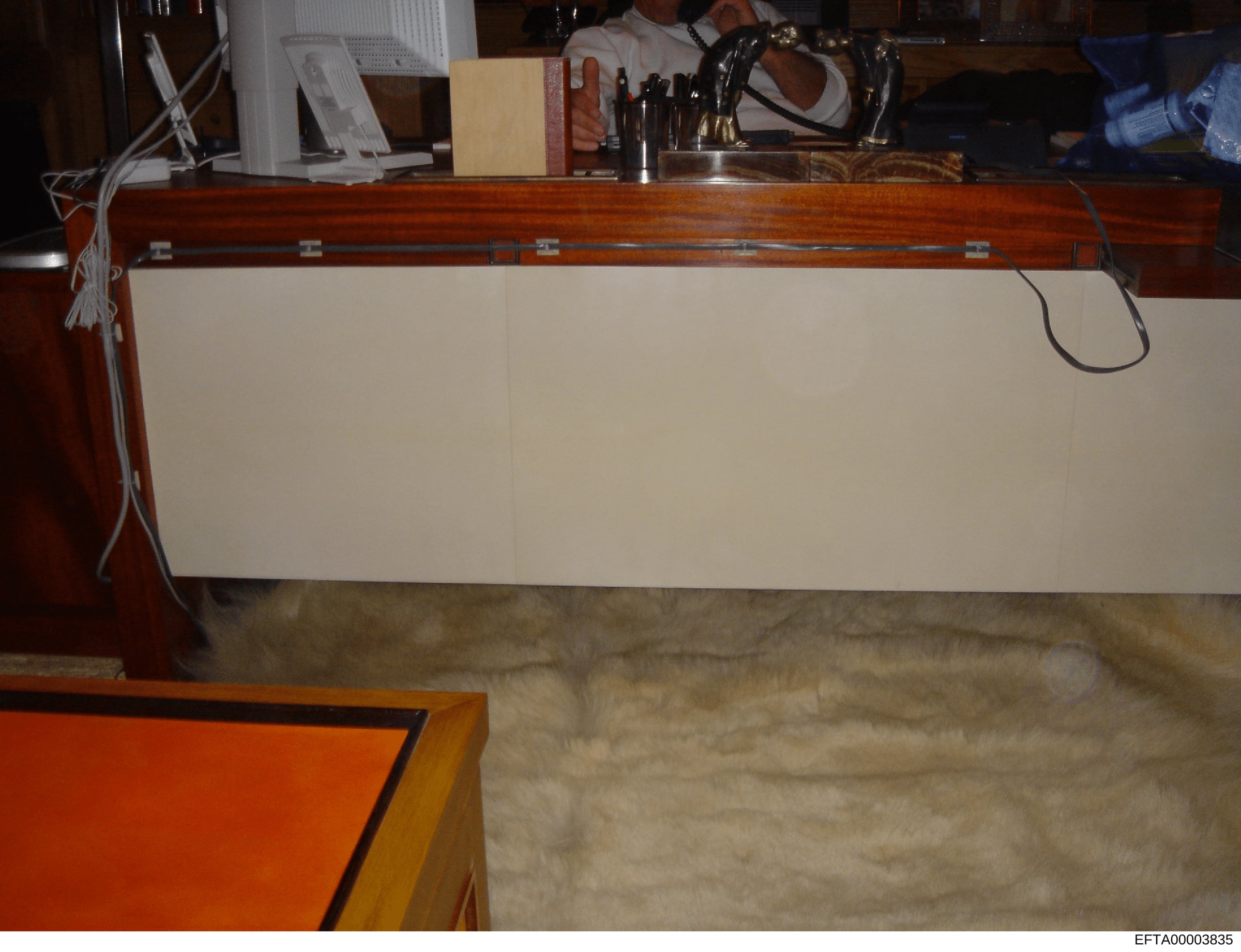 This is a photograph of an interior residential space showing a wooden desk or work surface with a cream-colored drawer front, various office items and personal effects on top, and what appears to be a carpeted floor. The image appears to be document